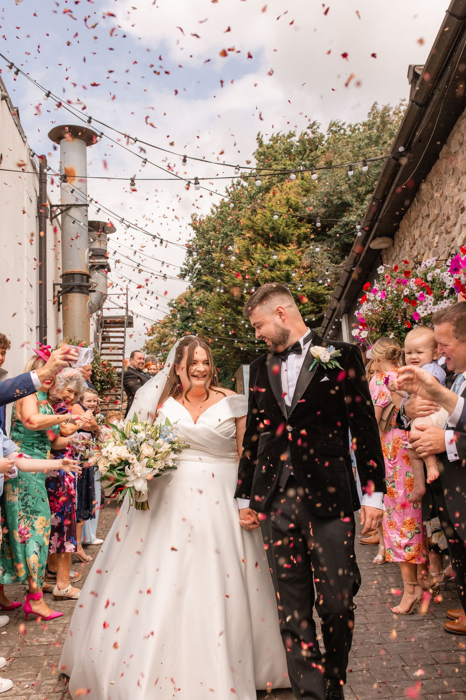 Wedding guests shower newlyweds with red confetti as they walk beneath string lights in a courtyard.
