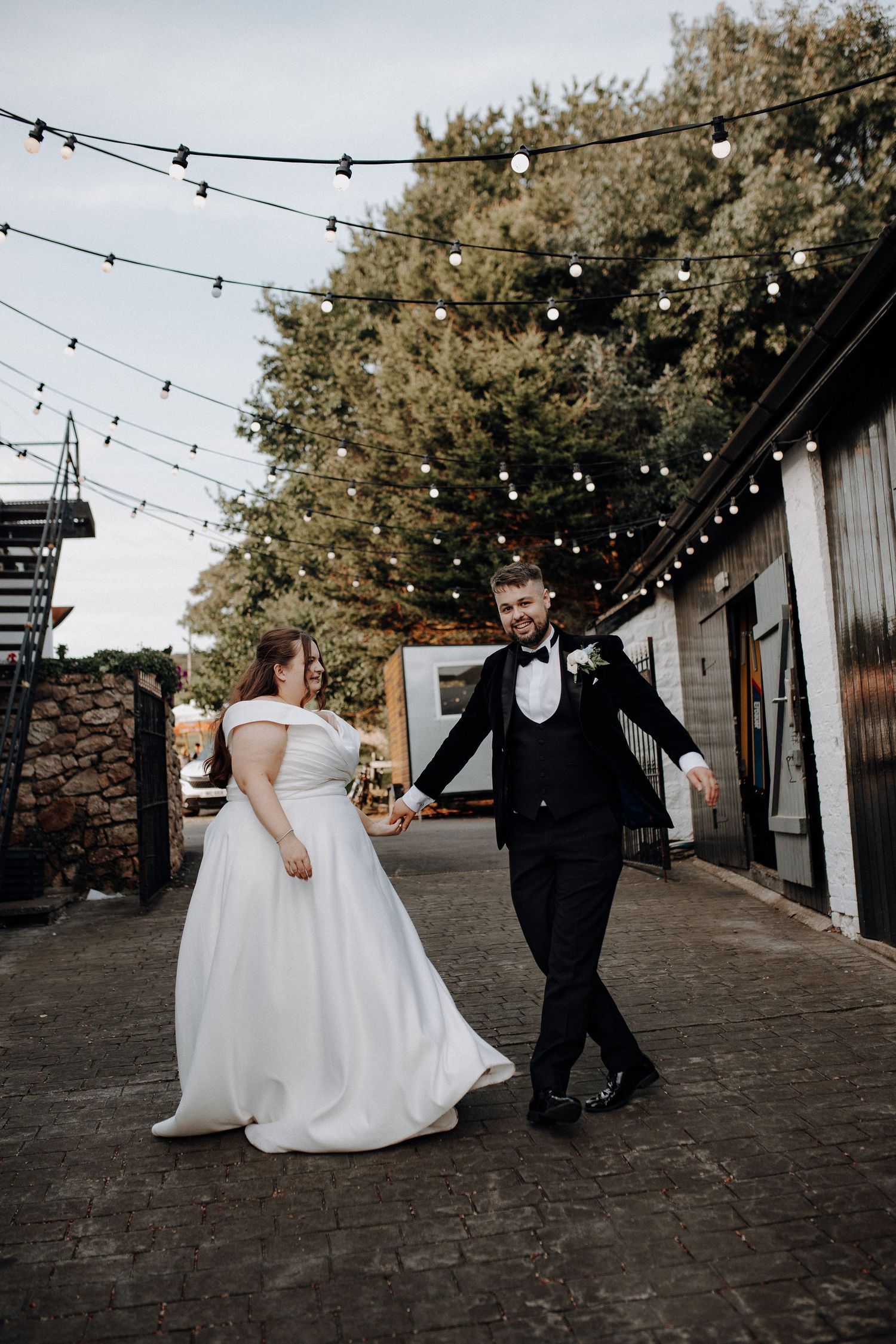 Wedding couple walking under string lights on cobblestone path at outdoor venue during sunset.
