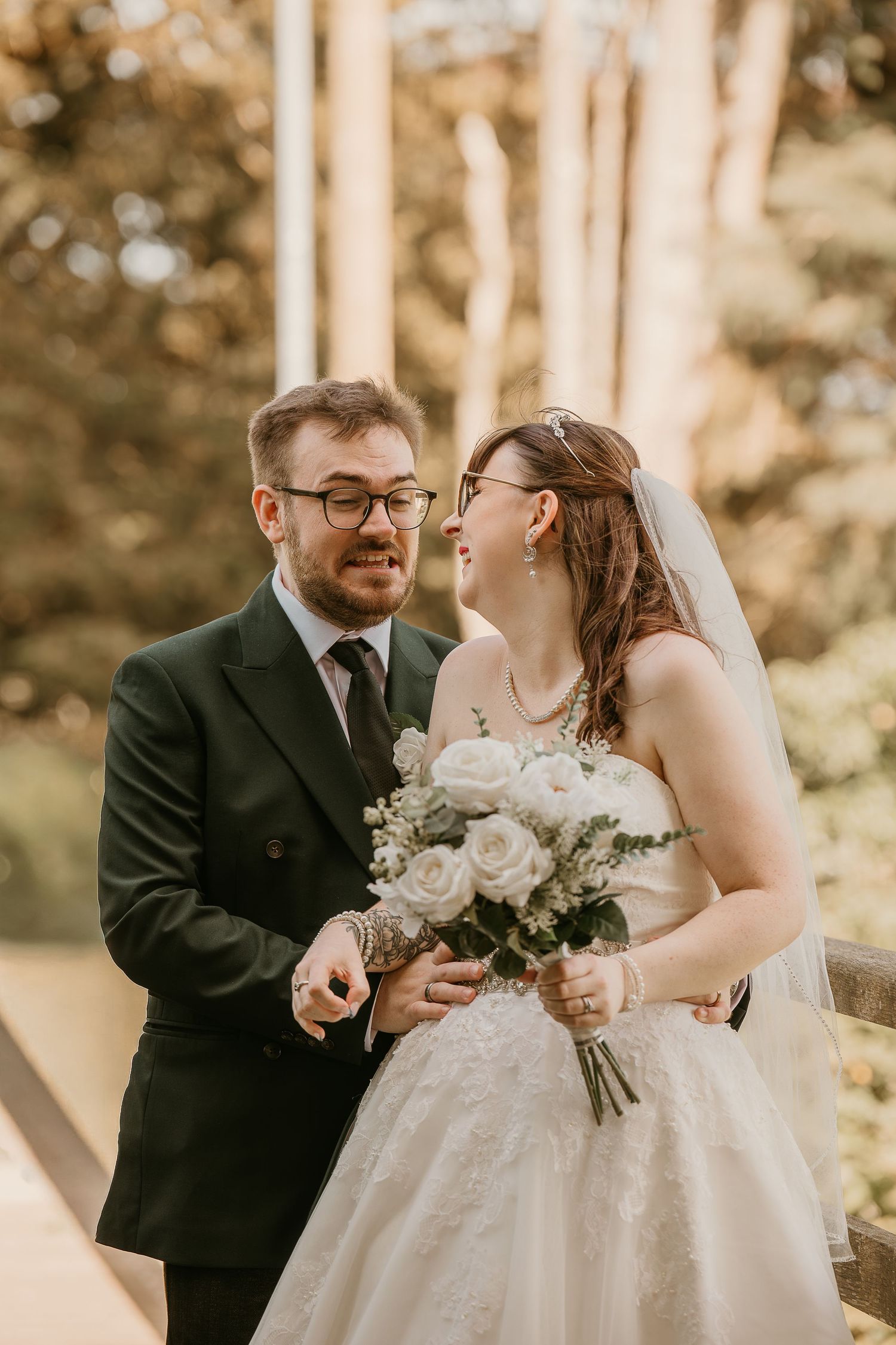 A bride and groom share a joyful moment outdoors, she holds white roses bouquet wearing a veil, he wears a dark suit.