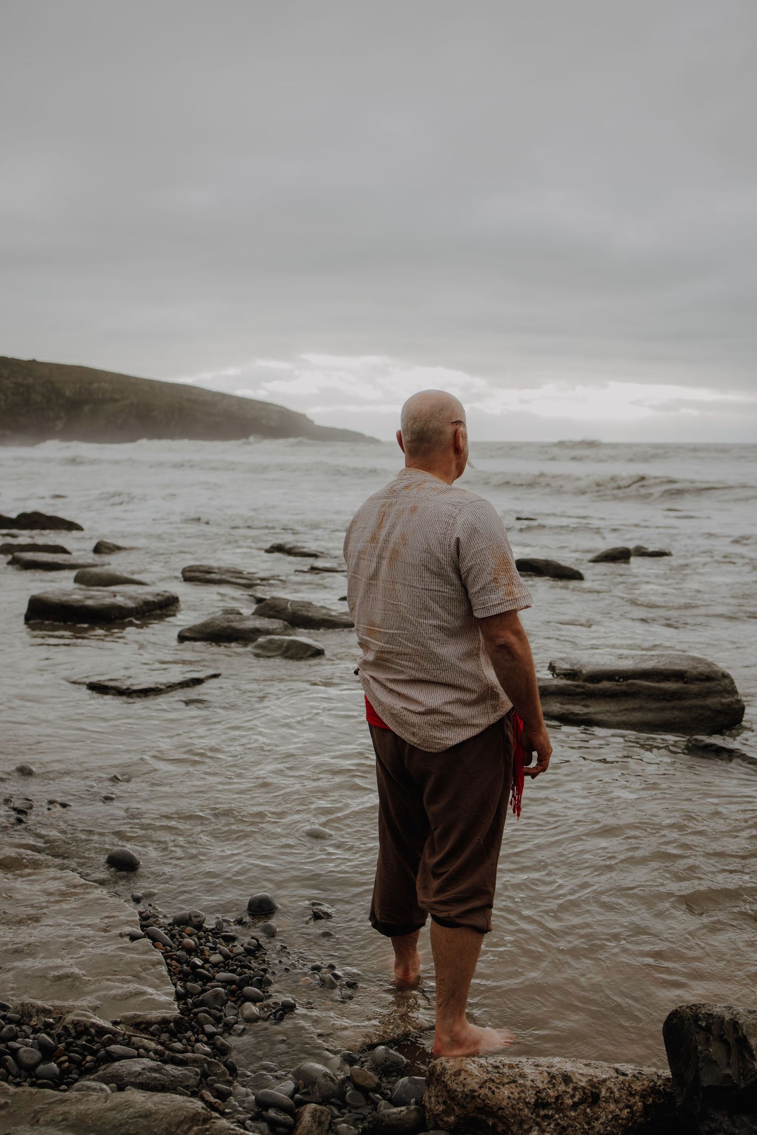 Person standing on rocky shore looking out at dramatic ocean waves.