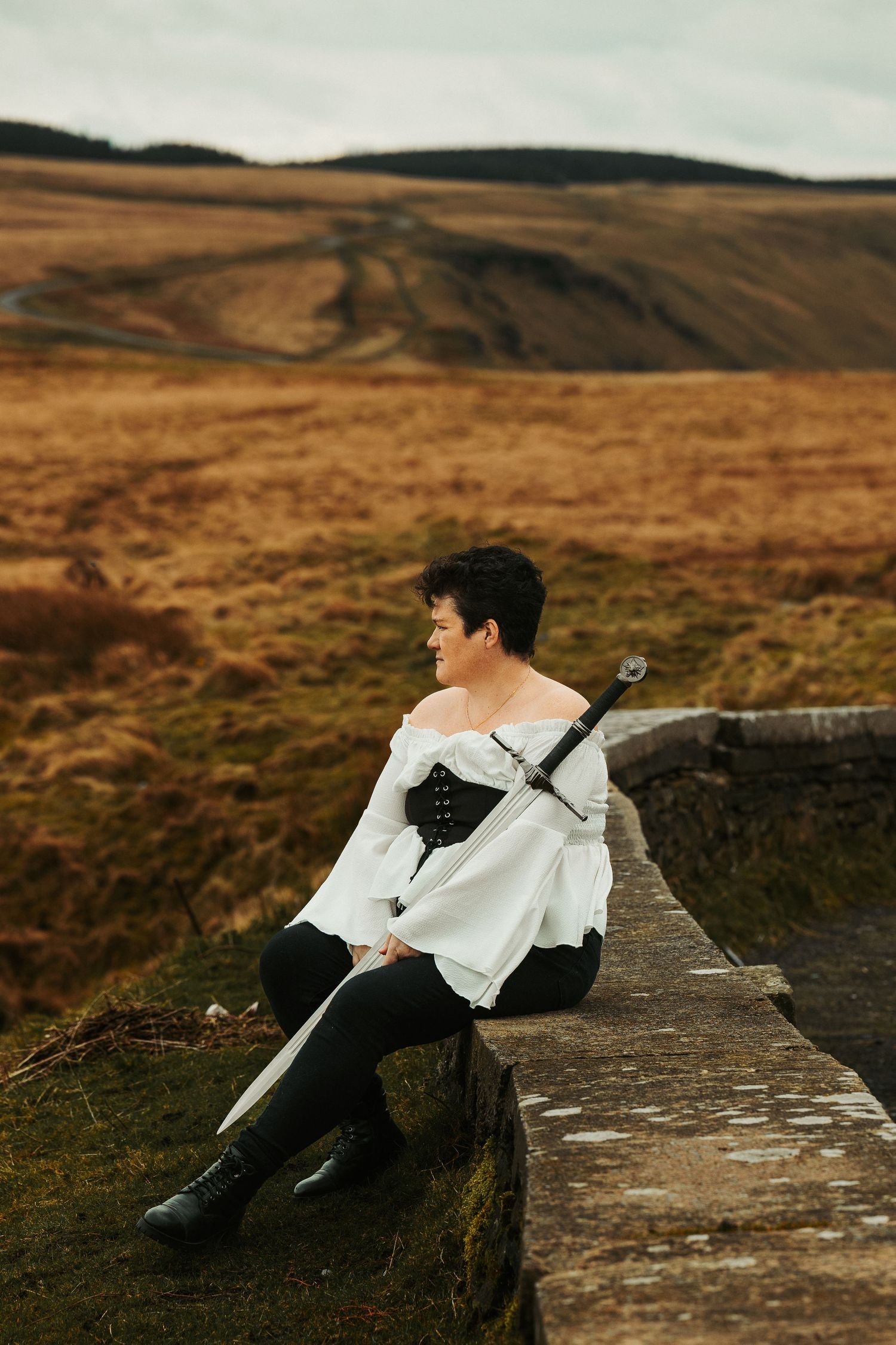 Figure in historical costume sits on stone wall overlooking scenic mountain landscape.