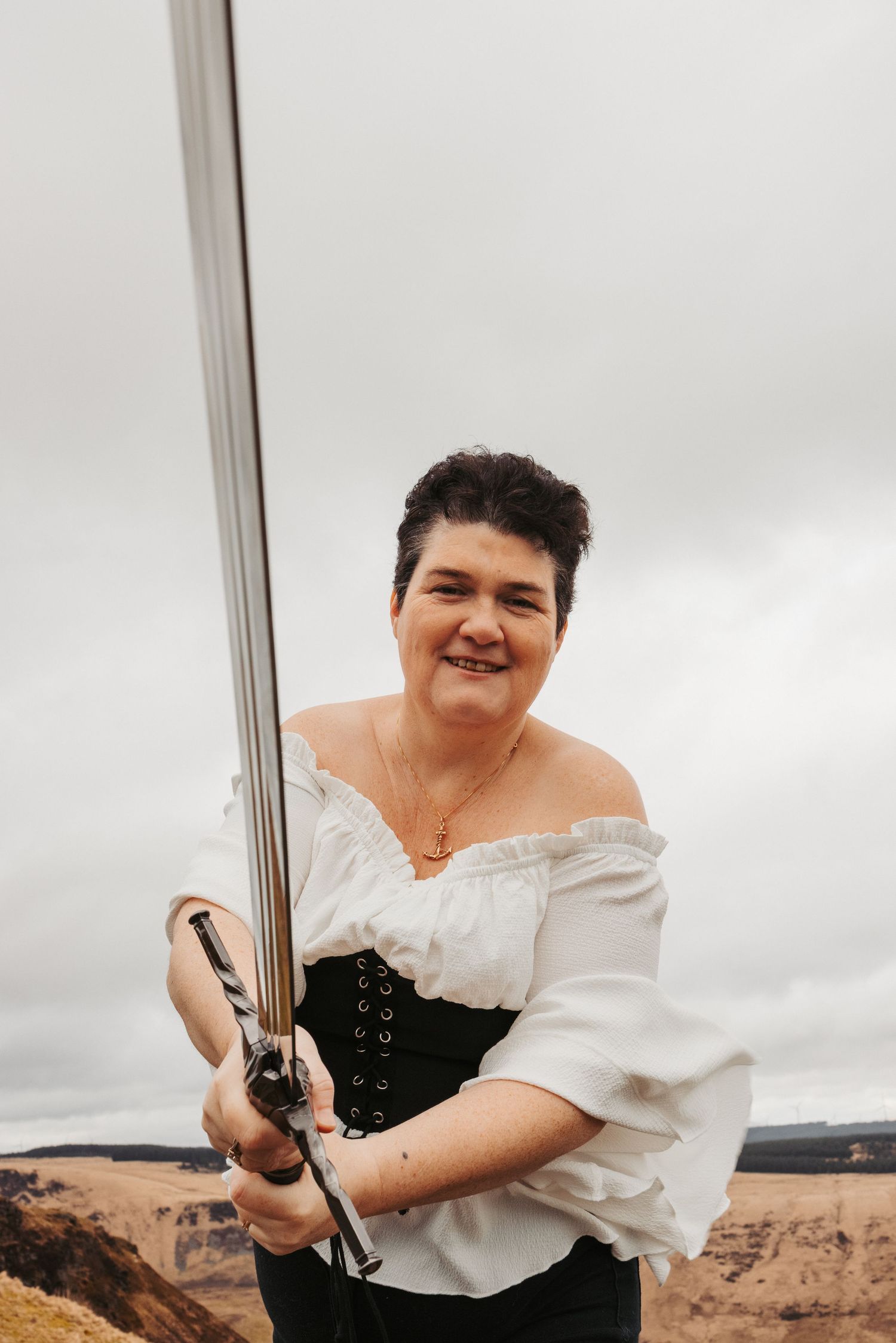 A sword wielder in a white peasant top and black corset poses against a cloudy sky.