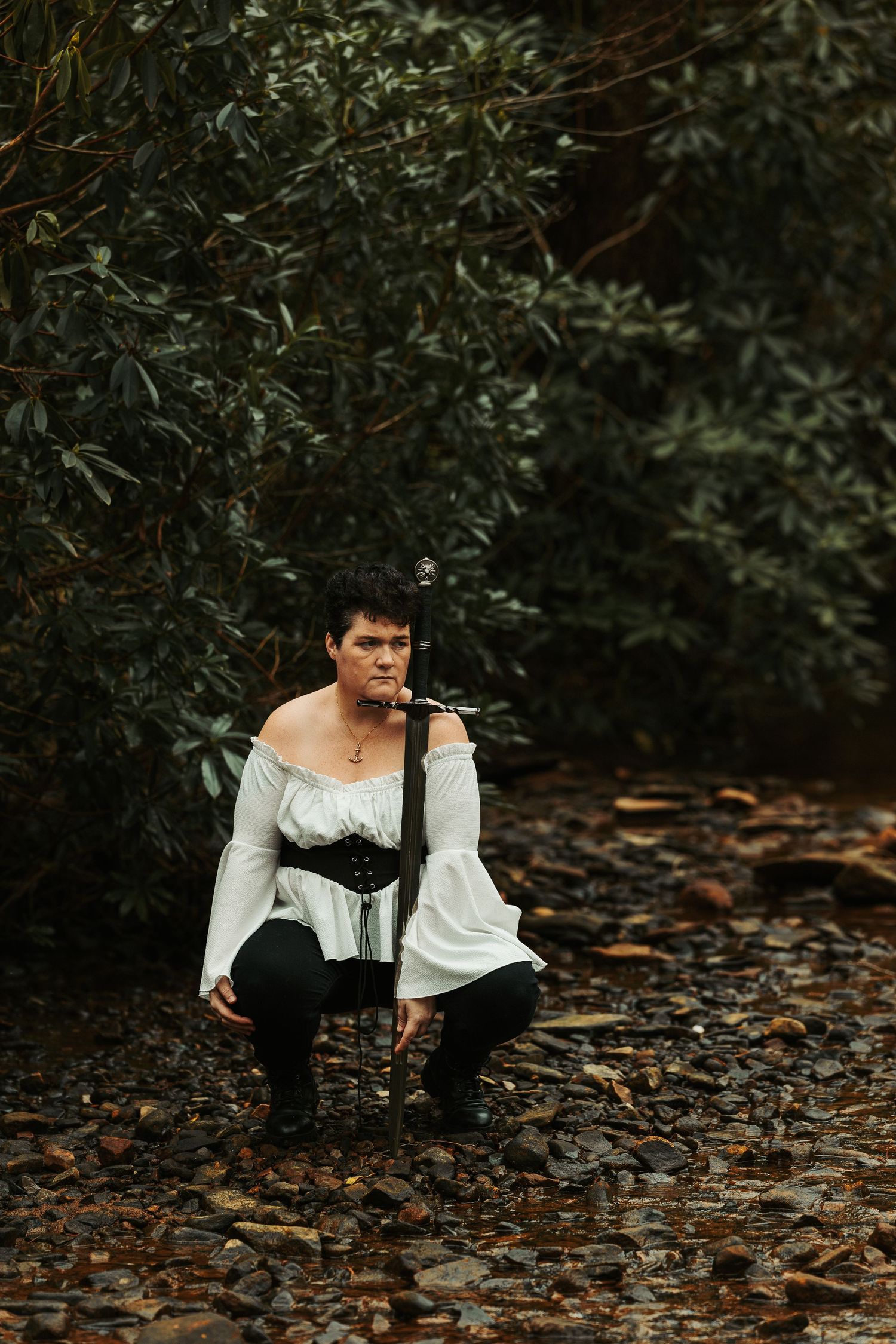 A figure in a white peasant blouse and black corset poses dramatically on fallen leaves in a forest setting.