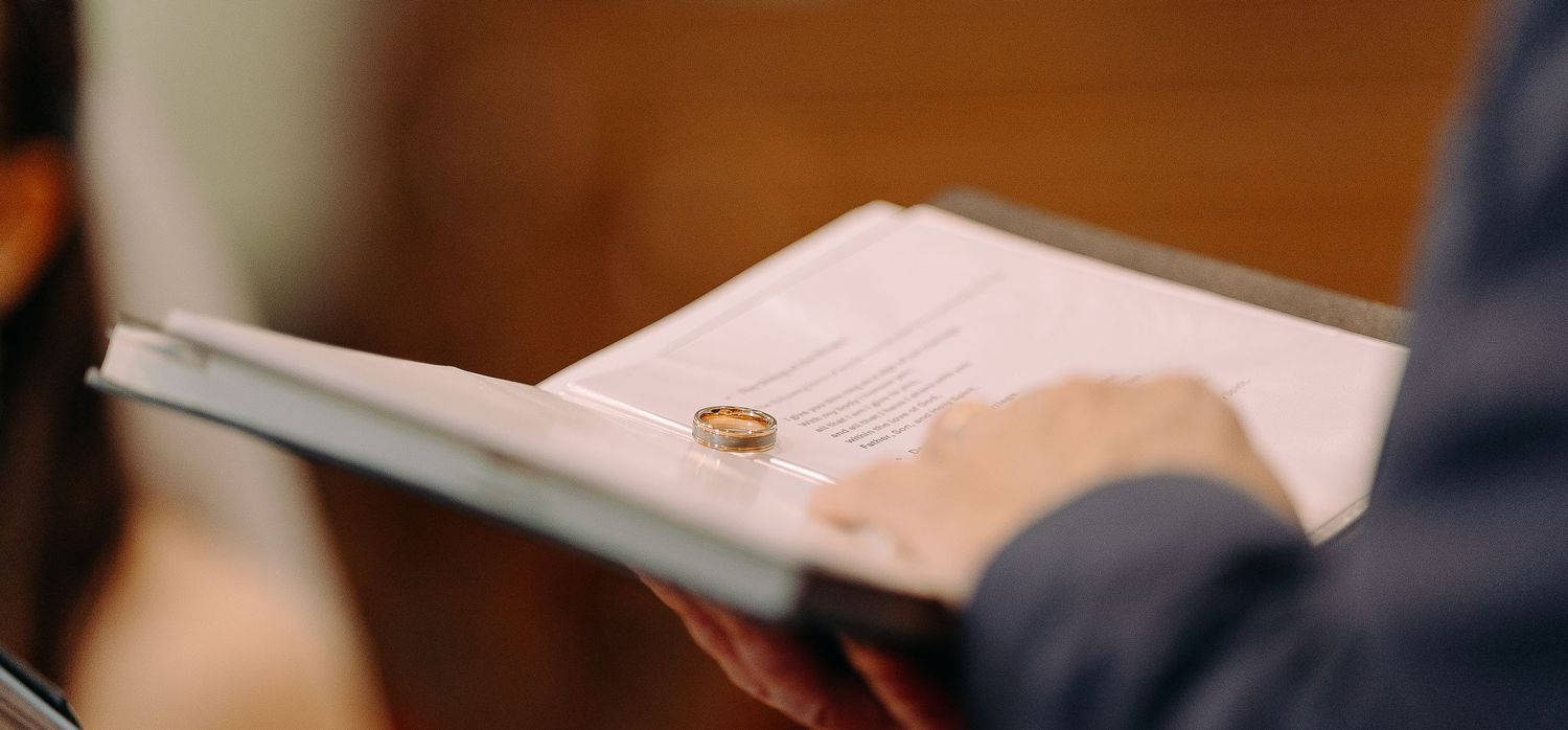 A gold wedding ring resting on an open book held by a person during a wedding ceremony.