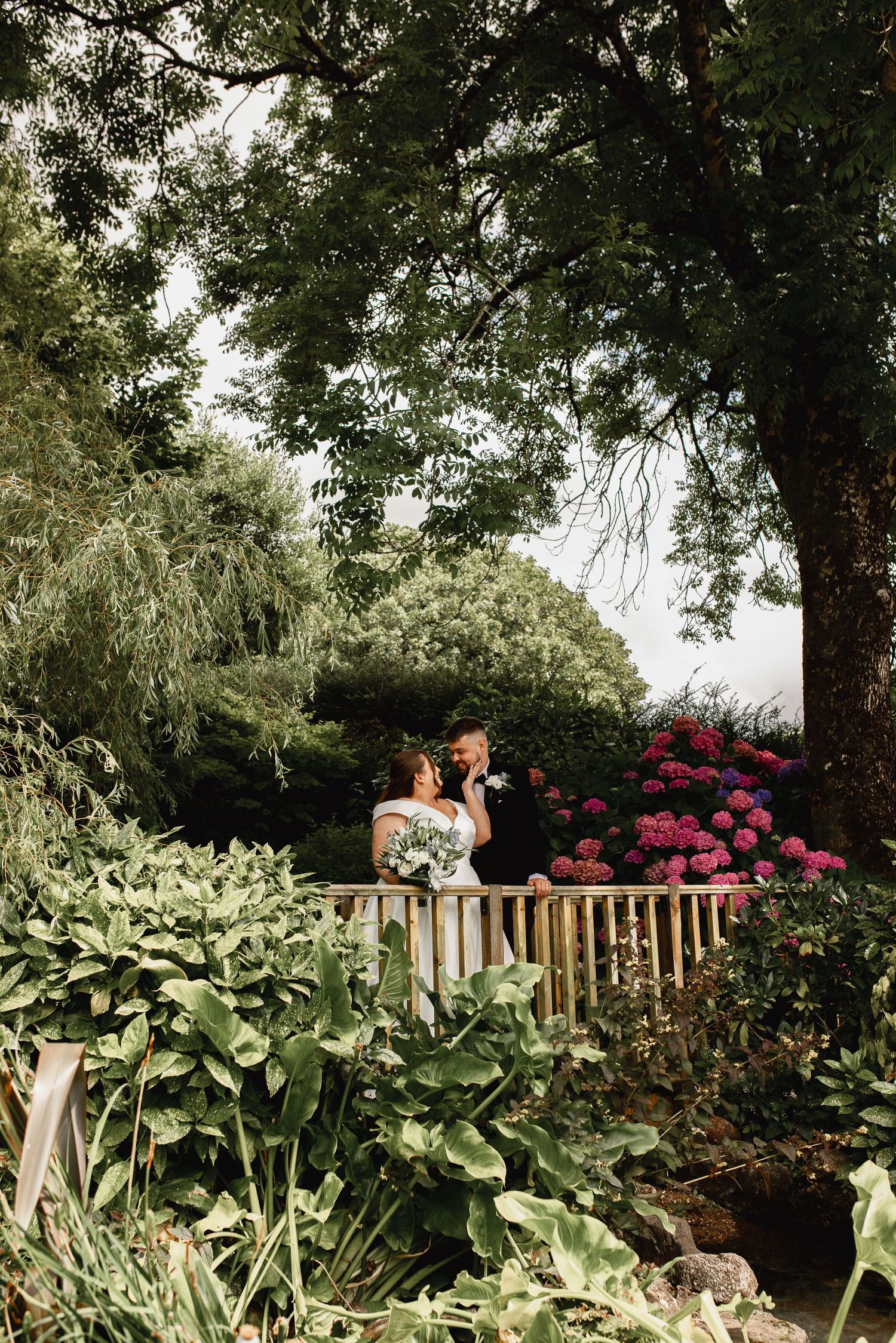 A couple stands on a wooden deck surrounded by lush garden foliage, blooming azaleas and towering trees.