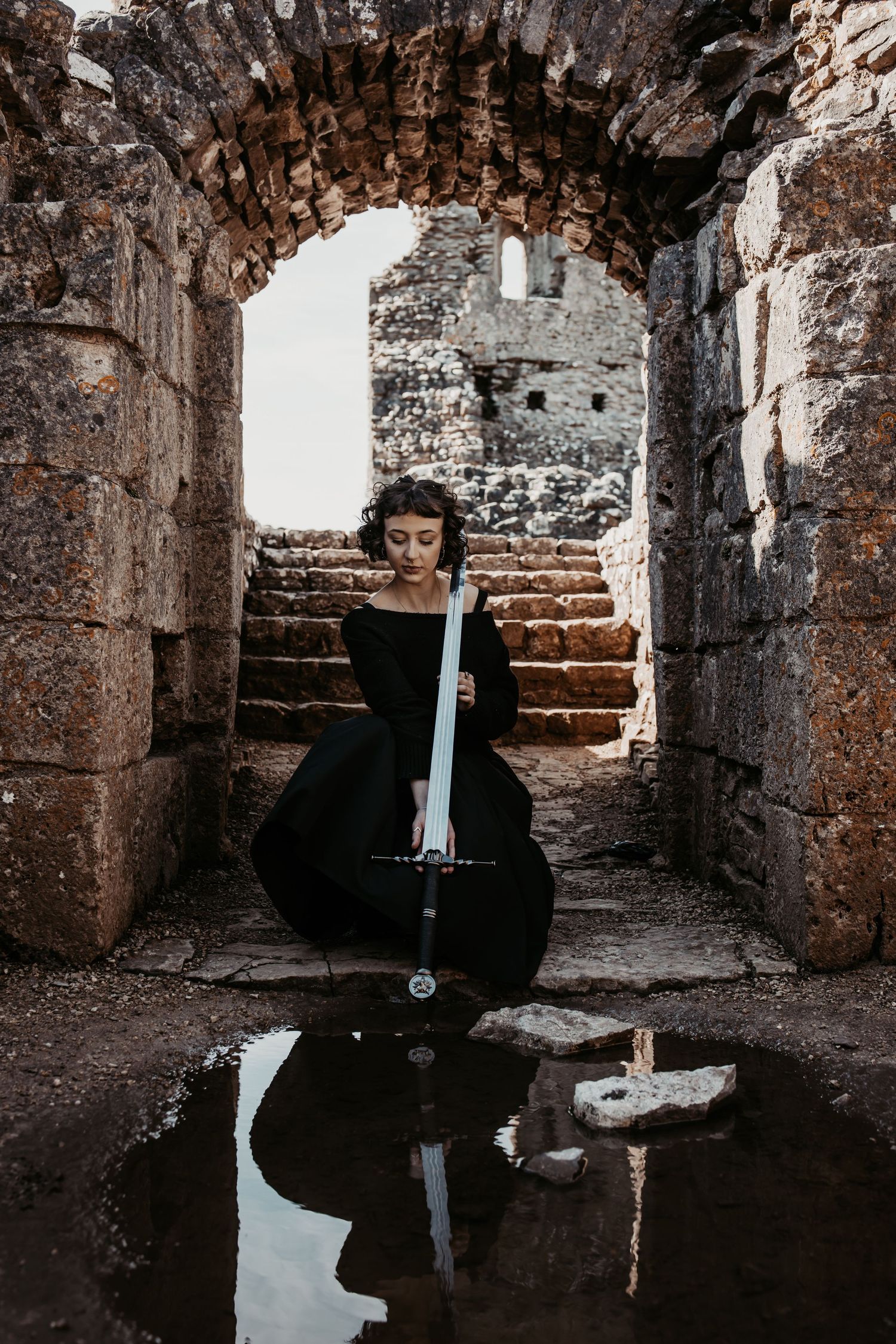 A person in black dress sits with sword in stone archway with reflection in puddle.