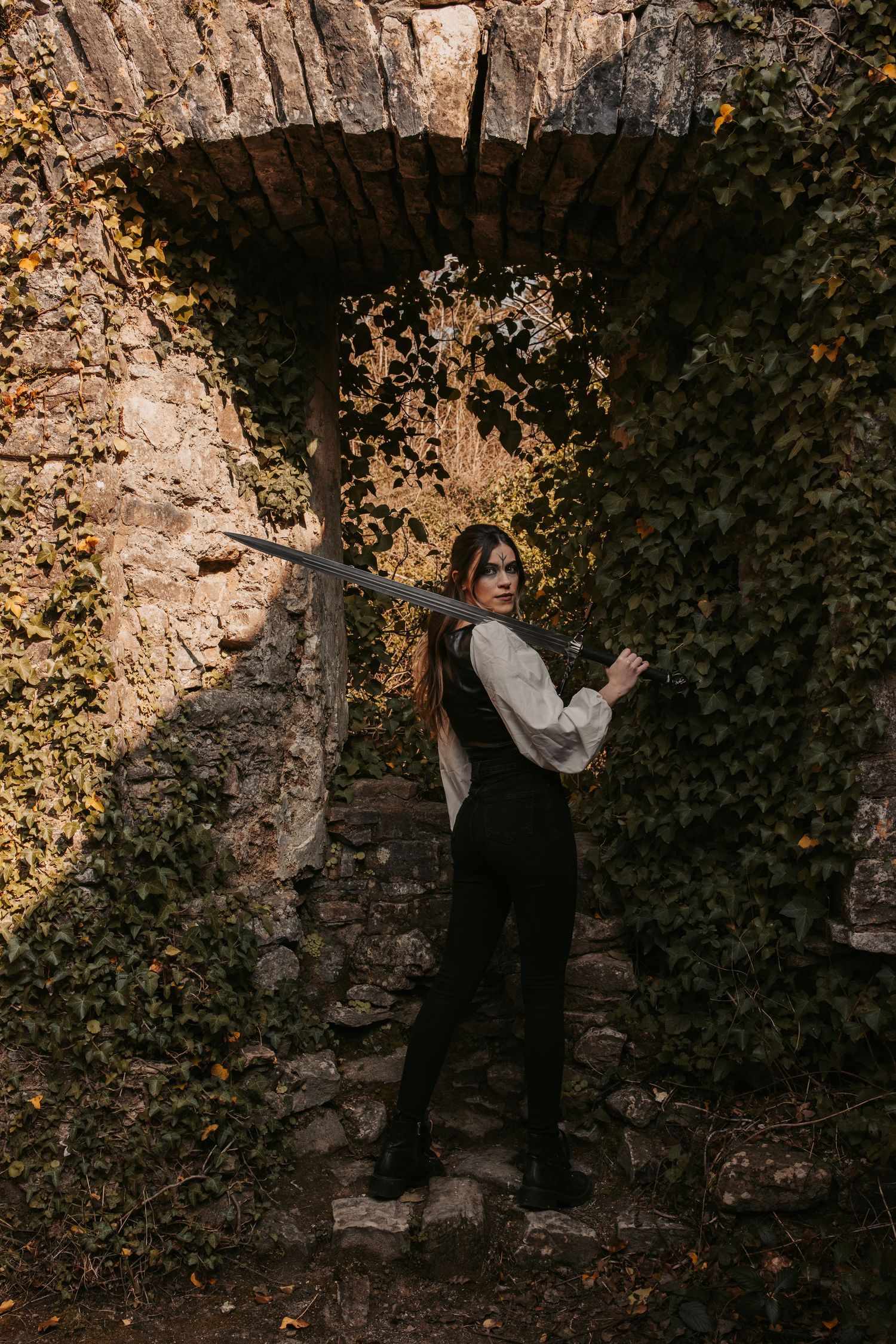 A figure in medieval dress poses with sword against an ivy-covered stone wall.