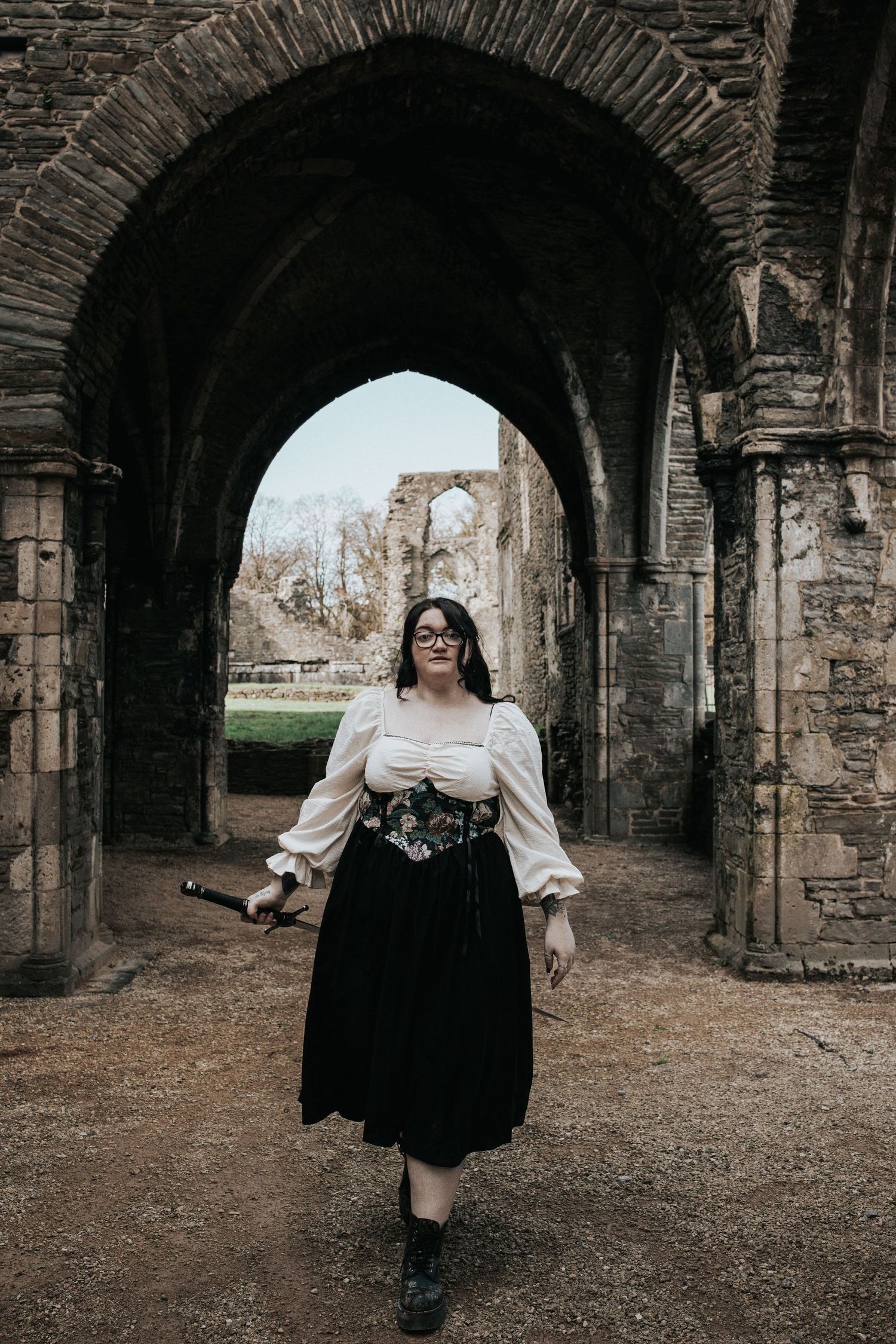 A figure in a white blouse and black dress walks through a stone archway corridor.
