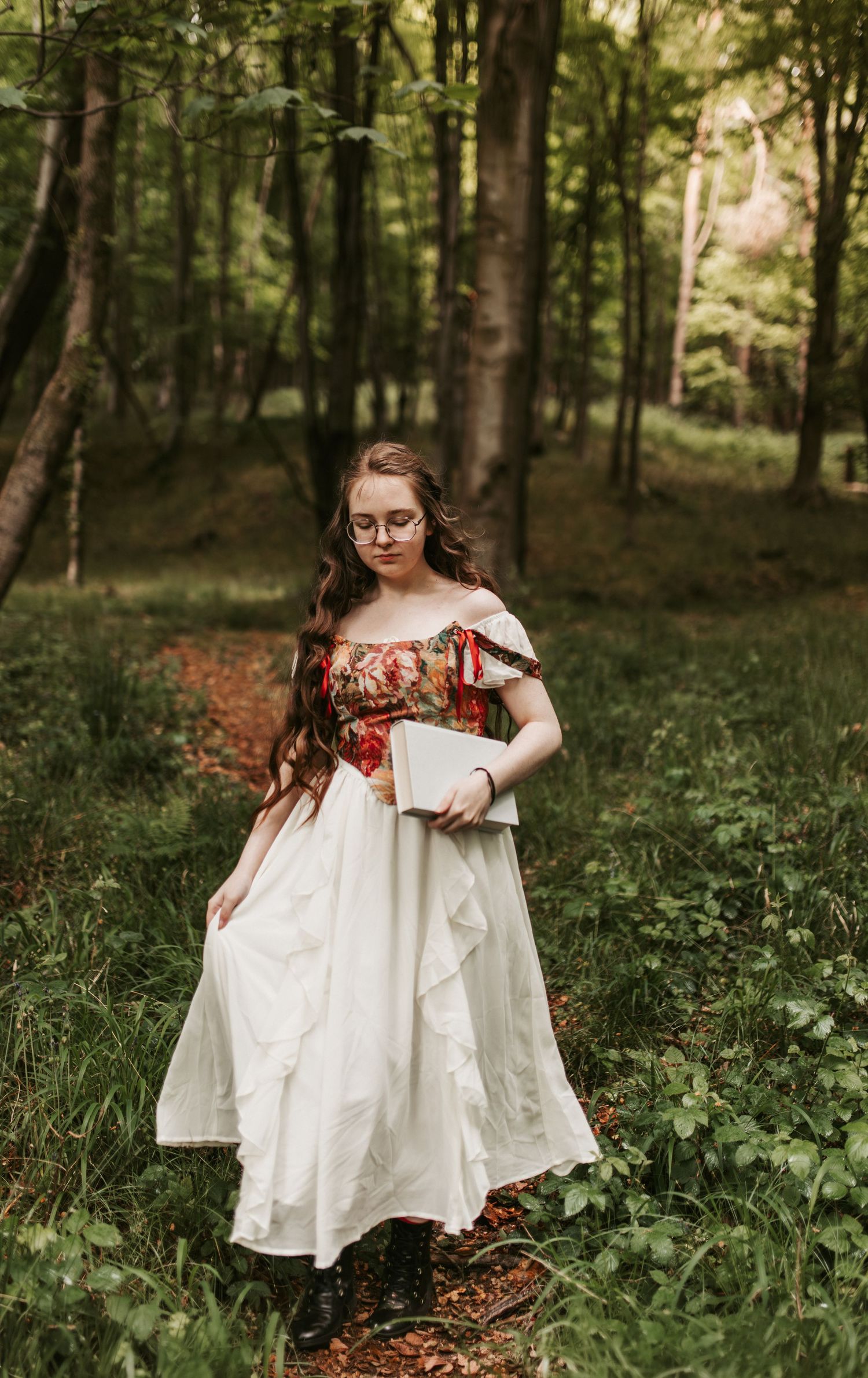Young woman in white floral Renaissance dress holding a book, standing in a lush green forest.