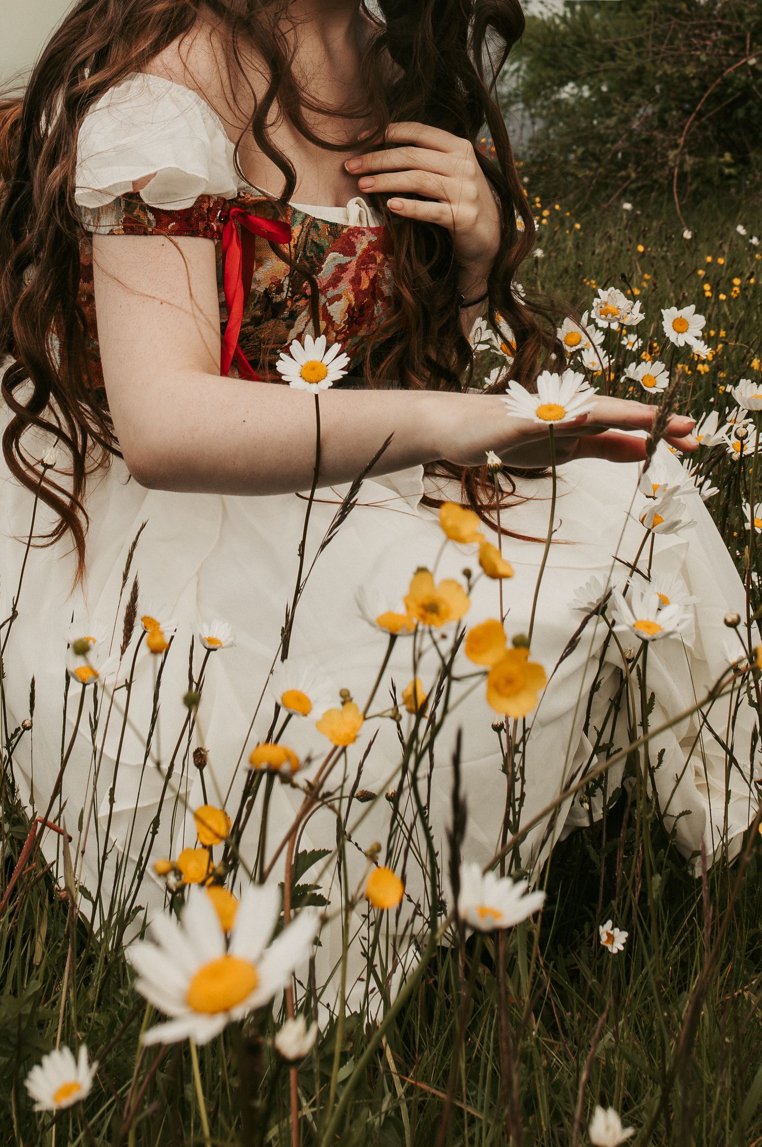 A young woman in a vintage white and red dress sits among wild daisies in a meadow, touching the flowers.