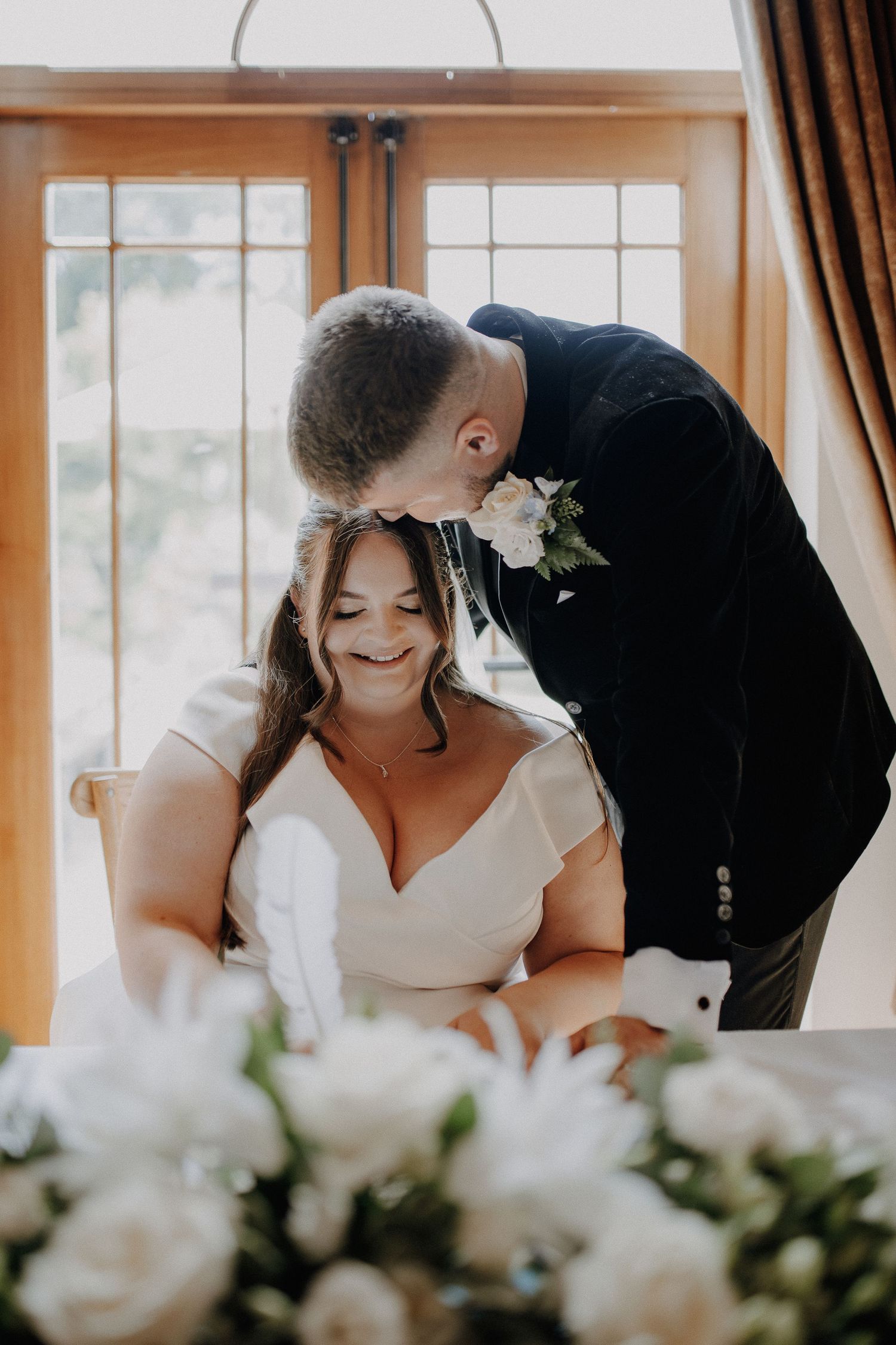 A tender wedding moment captured indoors with white floral decorations and warm lighting.