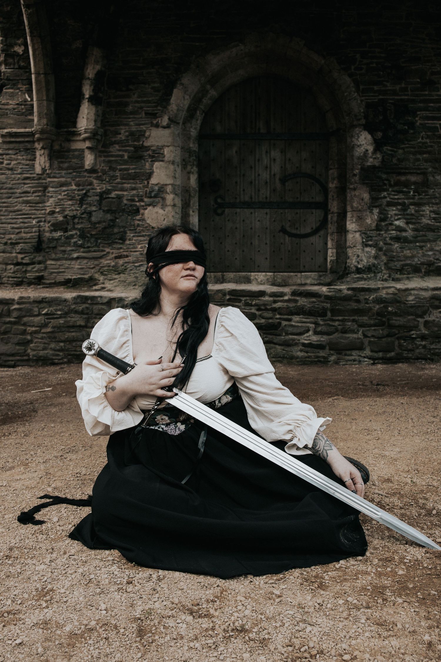 A person in historical costume reclines on ground with sword in gothic stone setting.