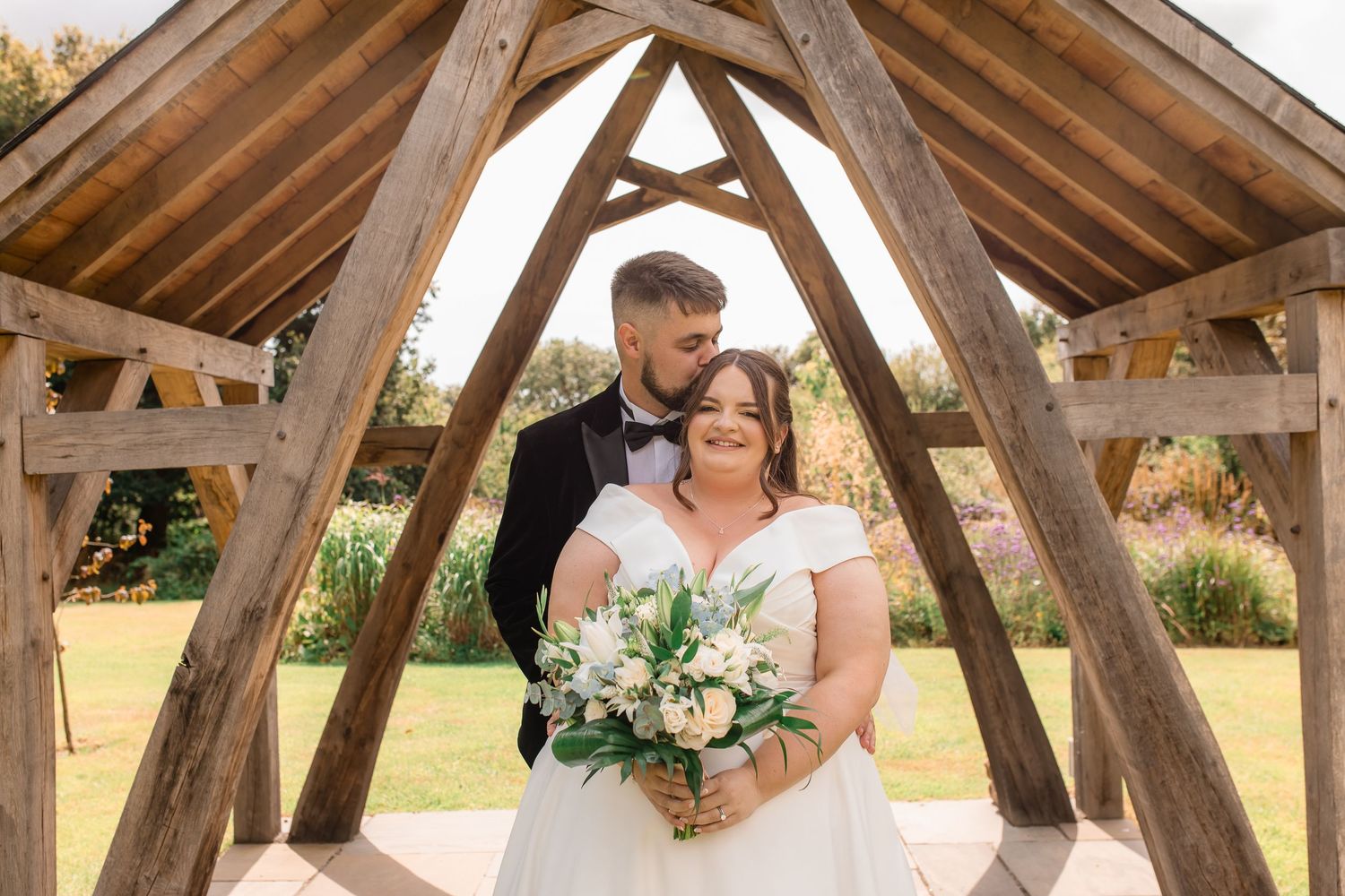 Wedding couple sharing intimate moments under a rustic wooden pavilion structure.