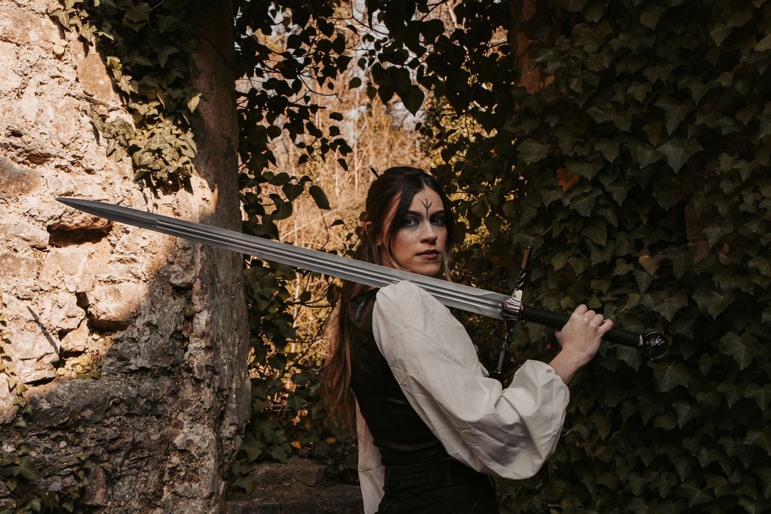 A figure in medieval attire holds a sword while standing near an ivy-covered wall.