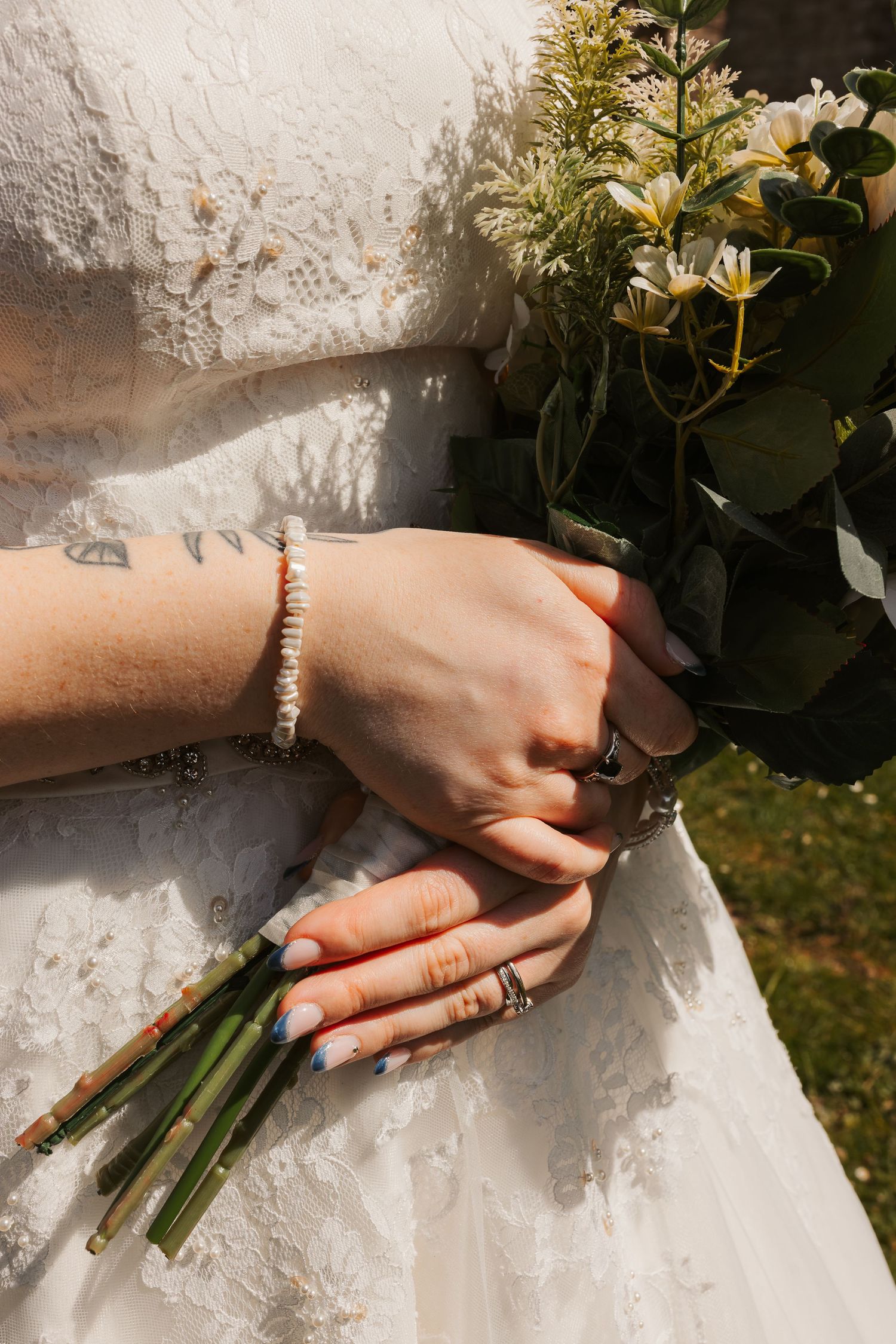 Bride holding a wildflower bouquet with lace wedding dress, pearl bracelet, rings, and French tip nails.