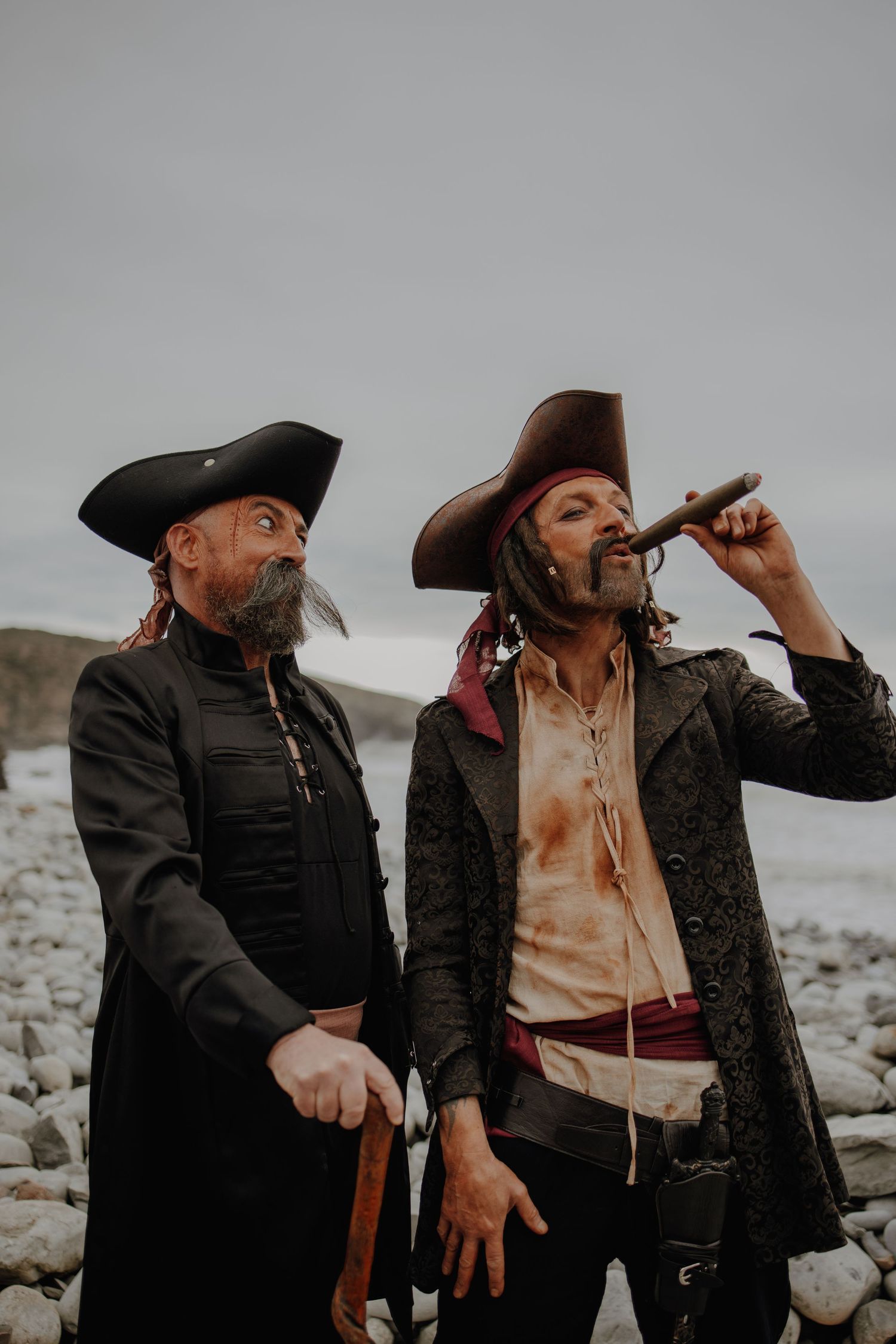 Two pirates in period costumes drinking from bottles on a rocky beach.