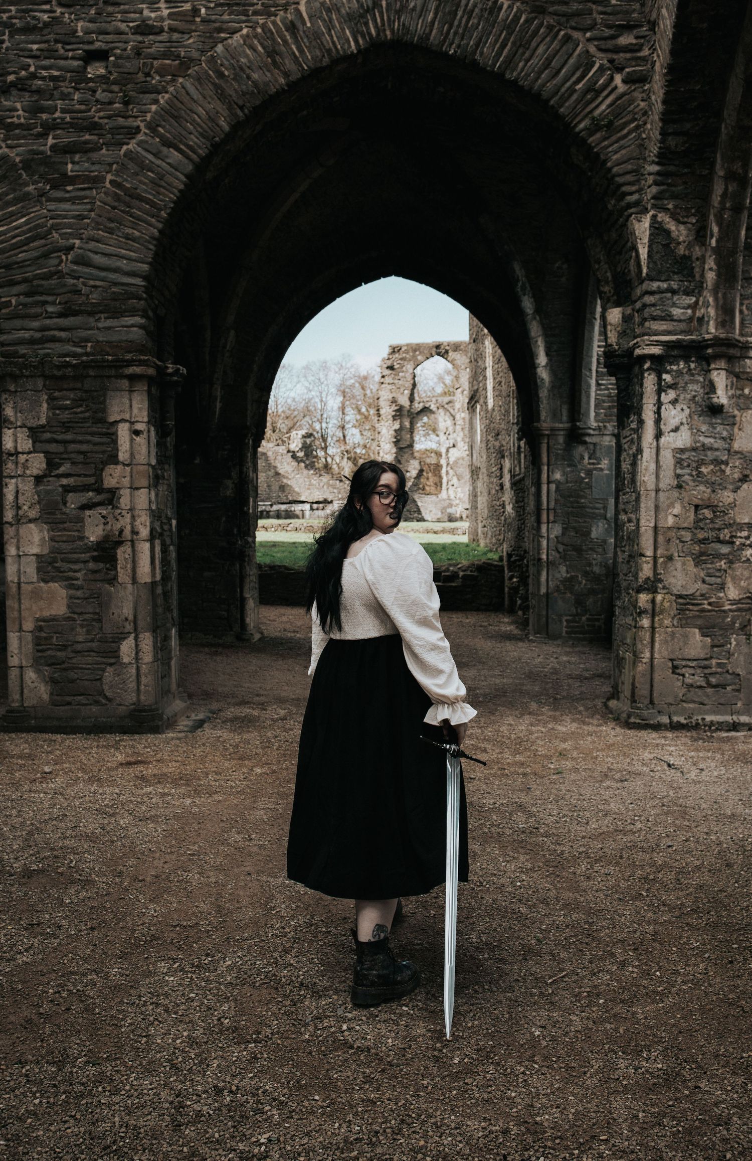 A person in historical costume poses with sword under stone arches of ancient ruins.