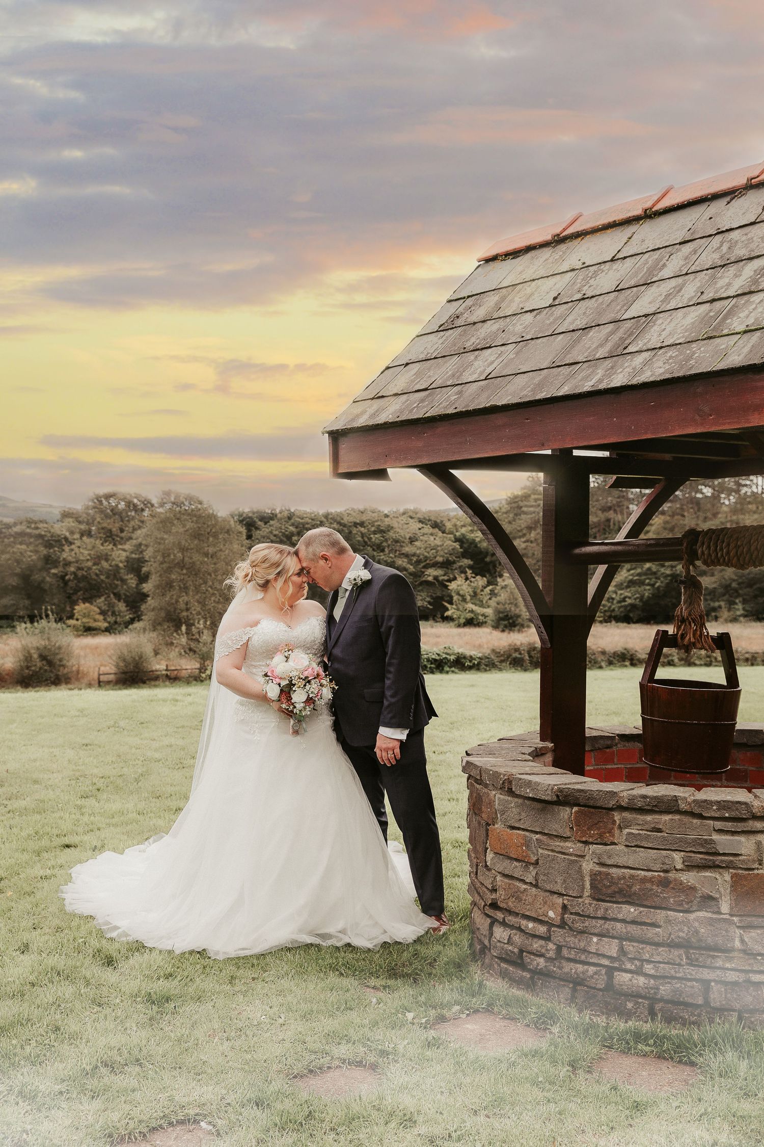 Romantic outdoor wedding photos taken at sunset by a rustic stone well with wooden shelter.