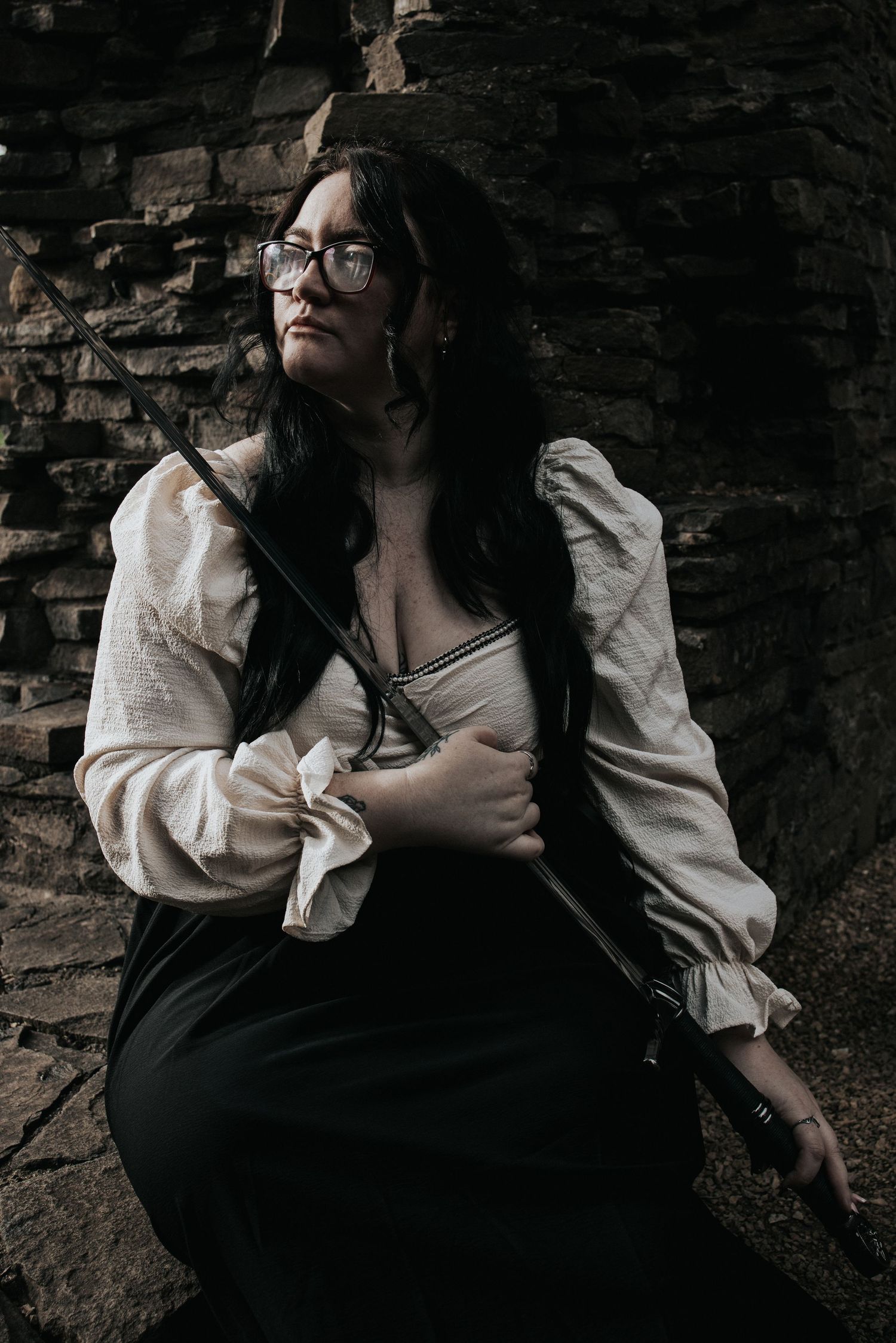 A dark-haired woman in glasses and a vintage white blouse with black skirt poses against a stone wall holding a sword.