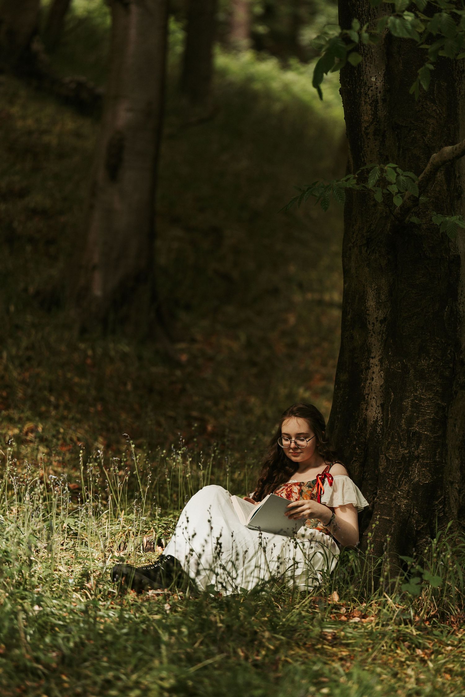 Young woman in white dress and glasses reading a book while sitting against a tree in a lush green forest.