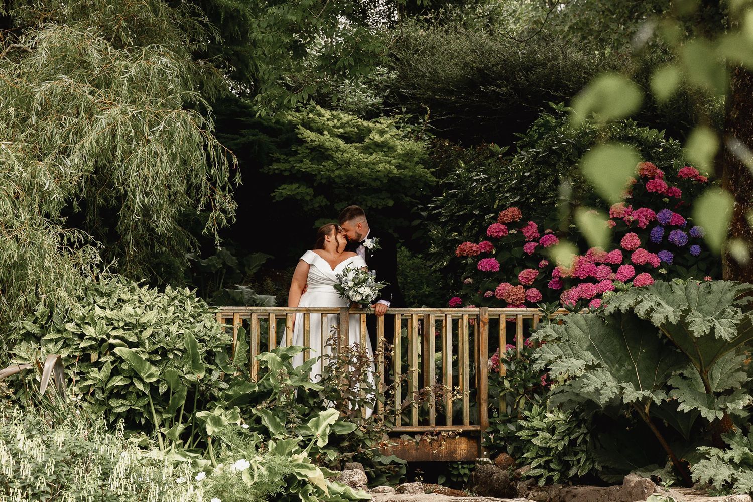 A romantic wedding couple embraces on a wooden bench surrounded by lush garden greenery and blooming flowers.