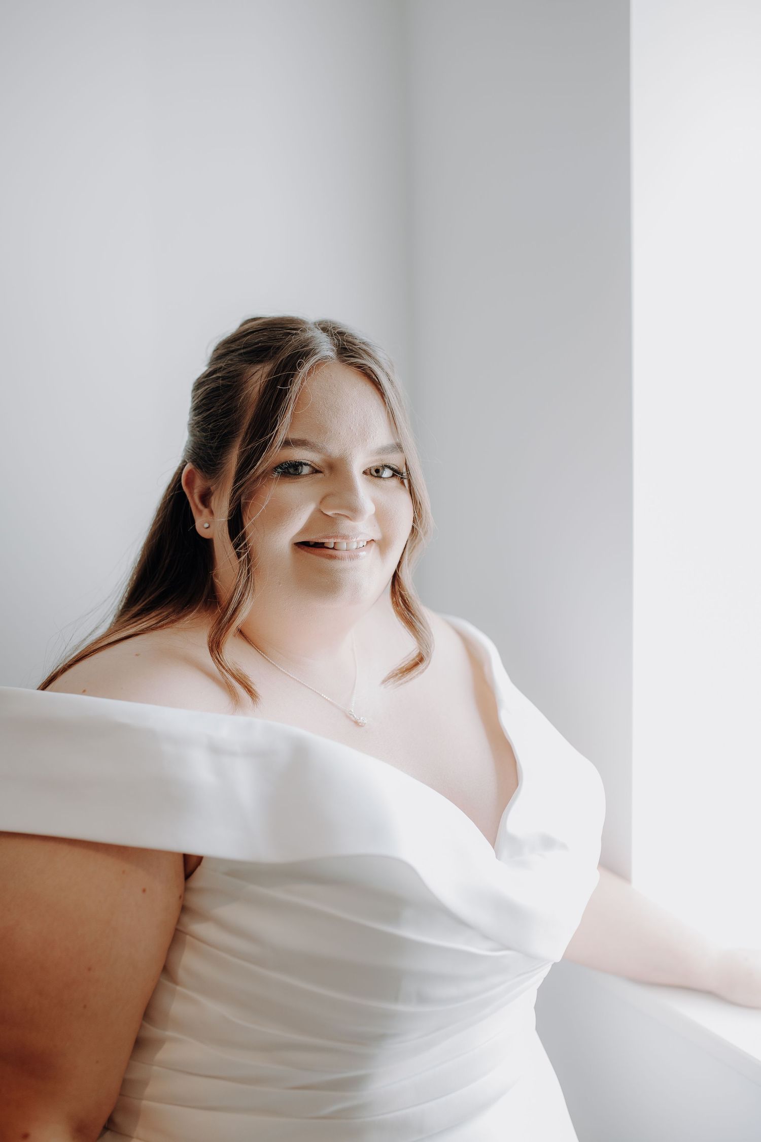 A bride in an elegant off-shoulder white wedding dress poses by a window with soft natural lighting.