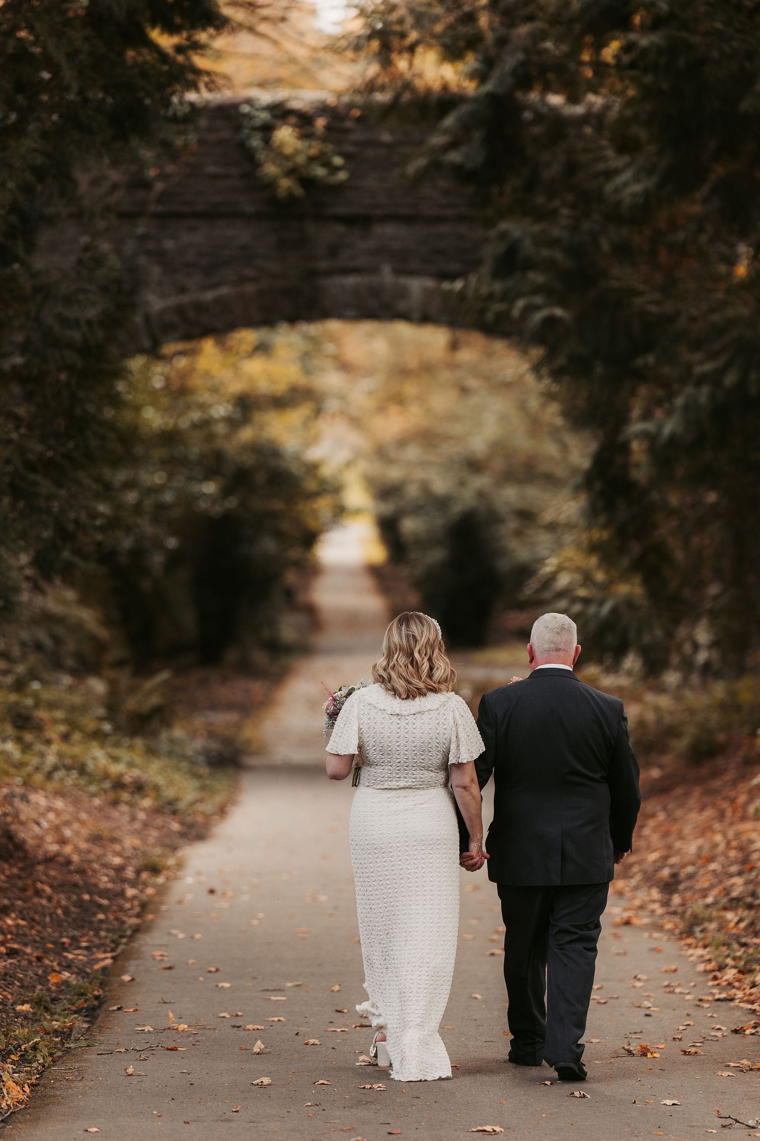 A couple in wedding attire walking down a tree-lined autumn path.