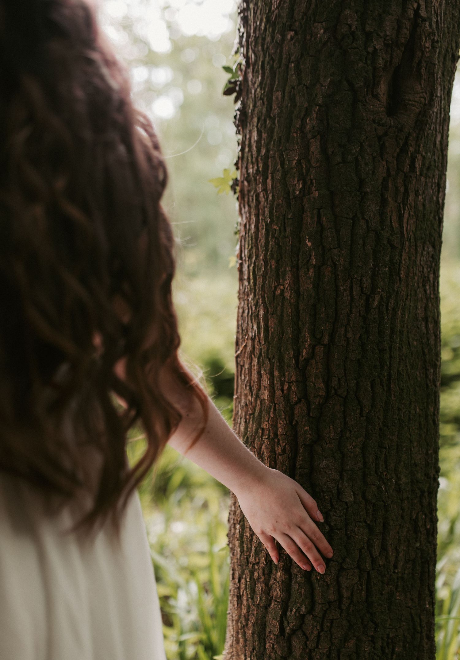 A young woman with curly brown hair in a white dress gently touches the bark of a large tree outdoors.