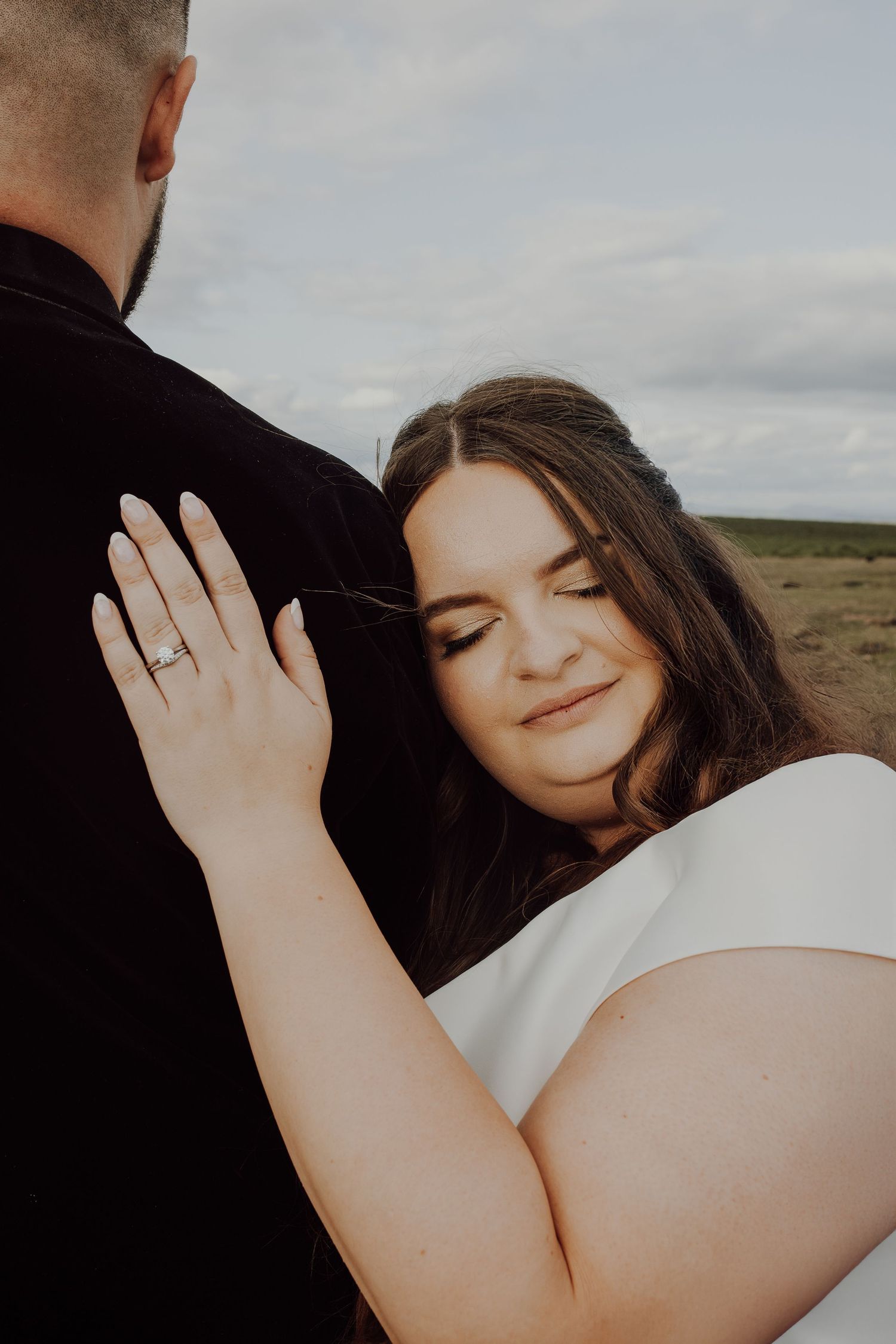 A romantic outdoor portrait of a couple embracing, with the bride wearing white and showing her engagement ring.