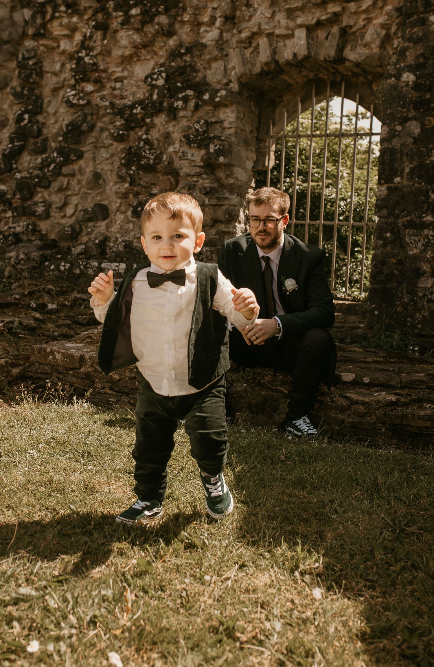 A young boy in a tuxedo and bow tie stands proudly outdoors while a man in a suit crouches behind him near stone ruins.