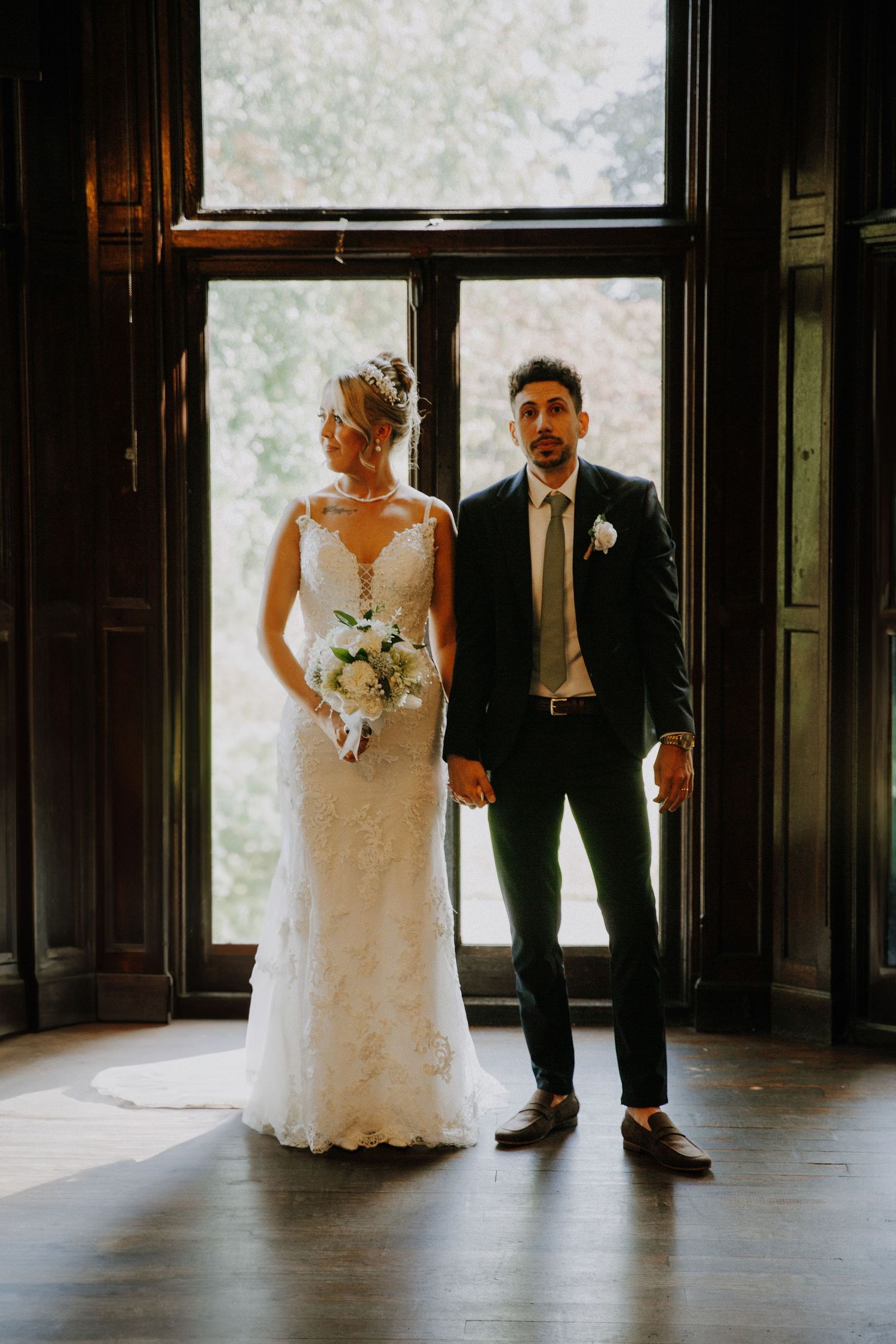 Wedding couple stands in elegant silhouette against bright windows in a dark interior space.