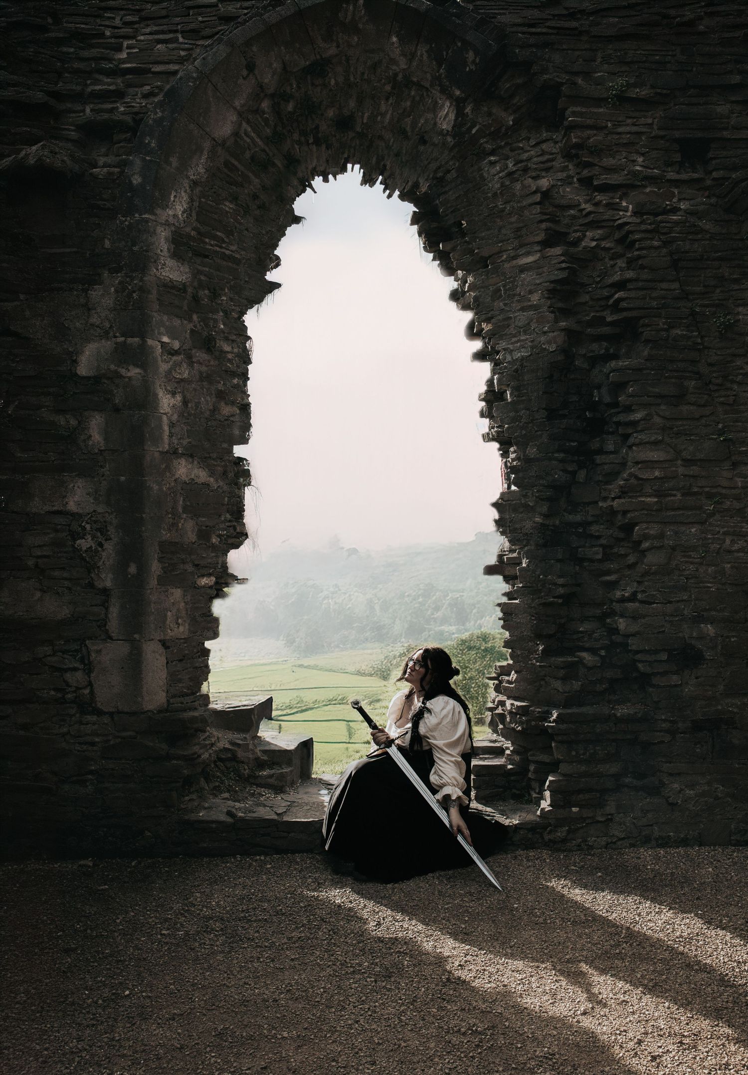 Silhouetted figure with sword sits in stone archway overlooking misty landscape.