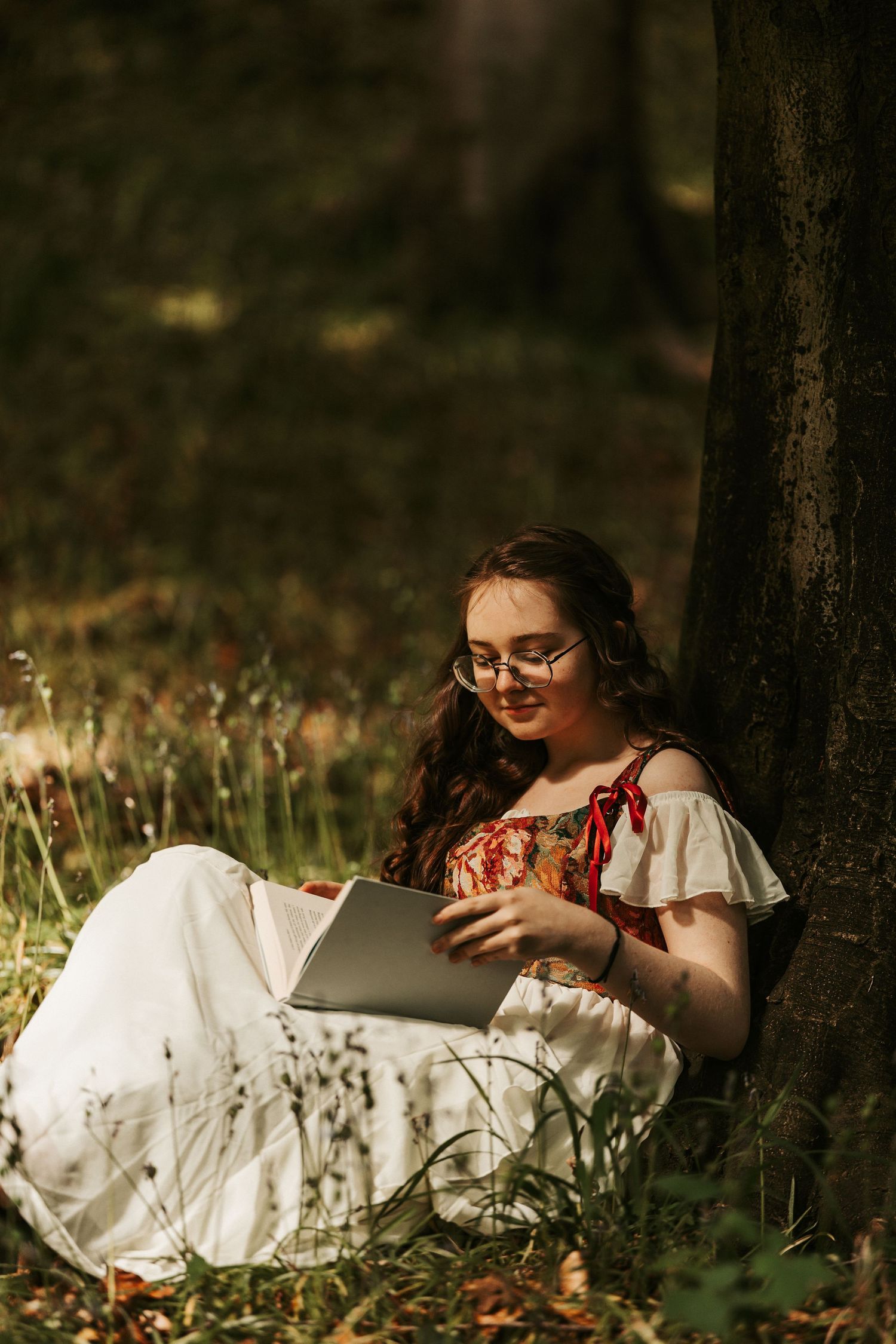Young woman in white floral dress reading a book while leaning against a tree in a sunlit forest.
