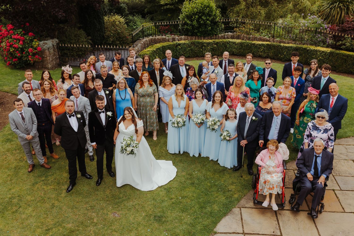 Large wedding party and guests gathered on a sunny lawn for outdoor group photos.