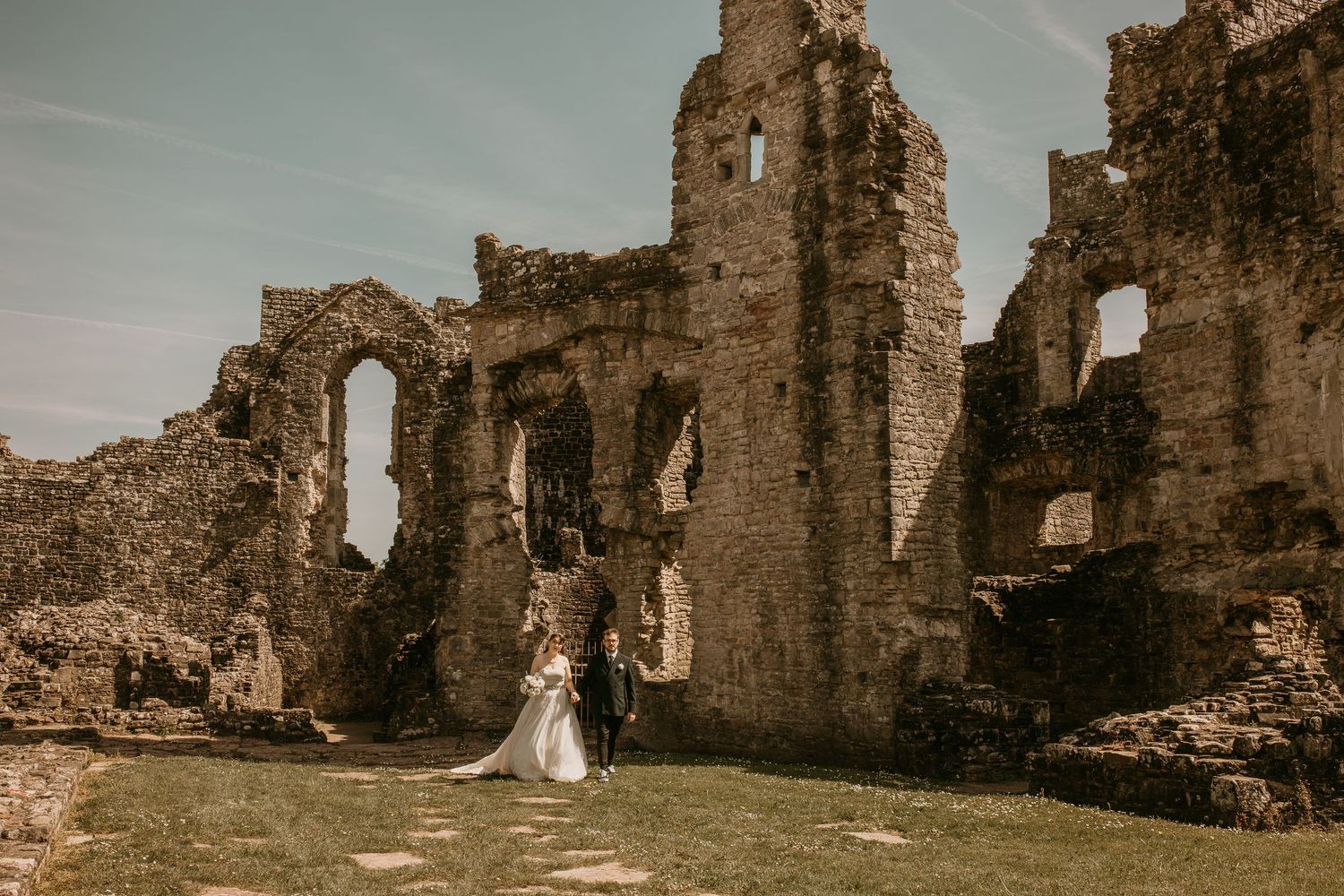 Bride and groom stand before ancient stone castle ruins on their wedding day under a blue sky.