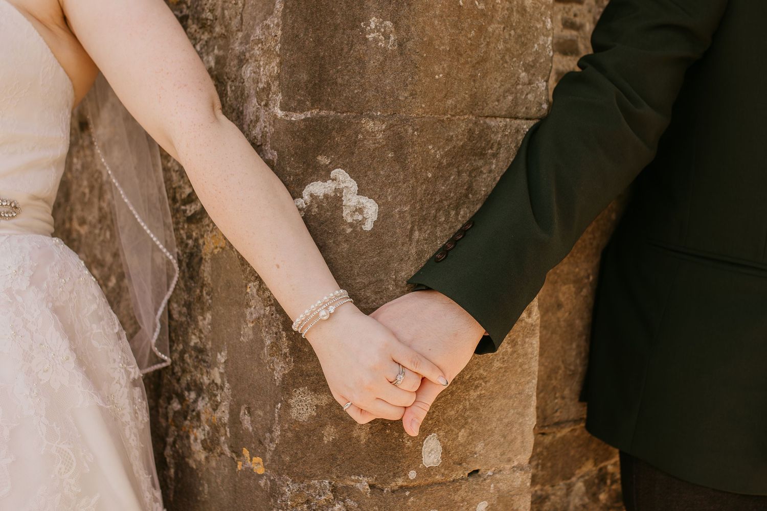 Close-up of bride and groom holding hands against a stone wall at their wedding.