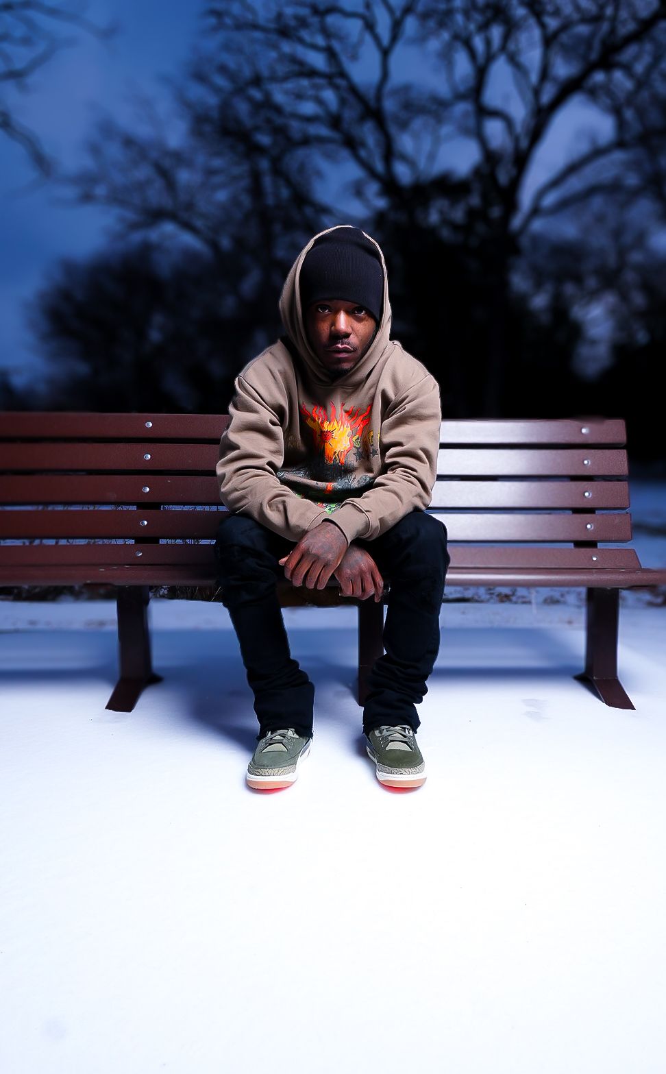 Person sitting alone on a wooden park bench at dusk wearing a light colored hoodie.