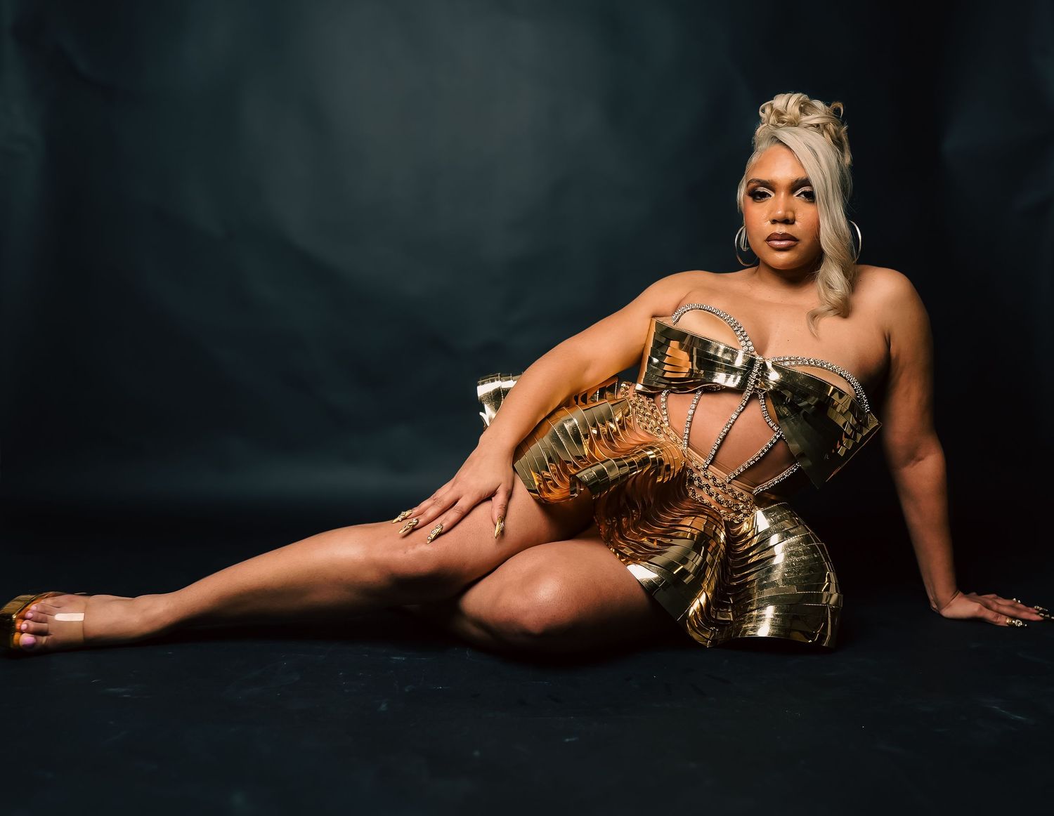 Woman posing in gold armored costume on dark studio floor, wearing blonde updo hairstyle.