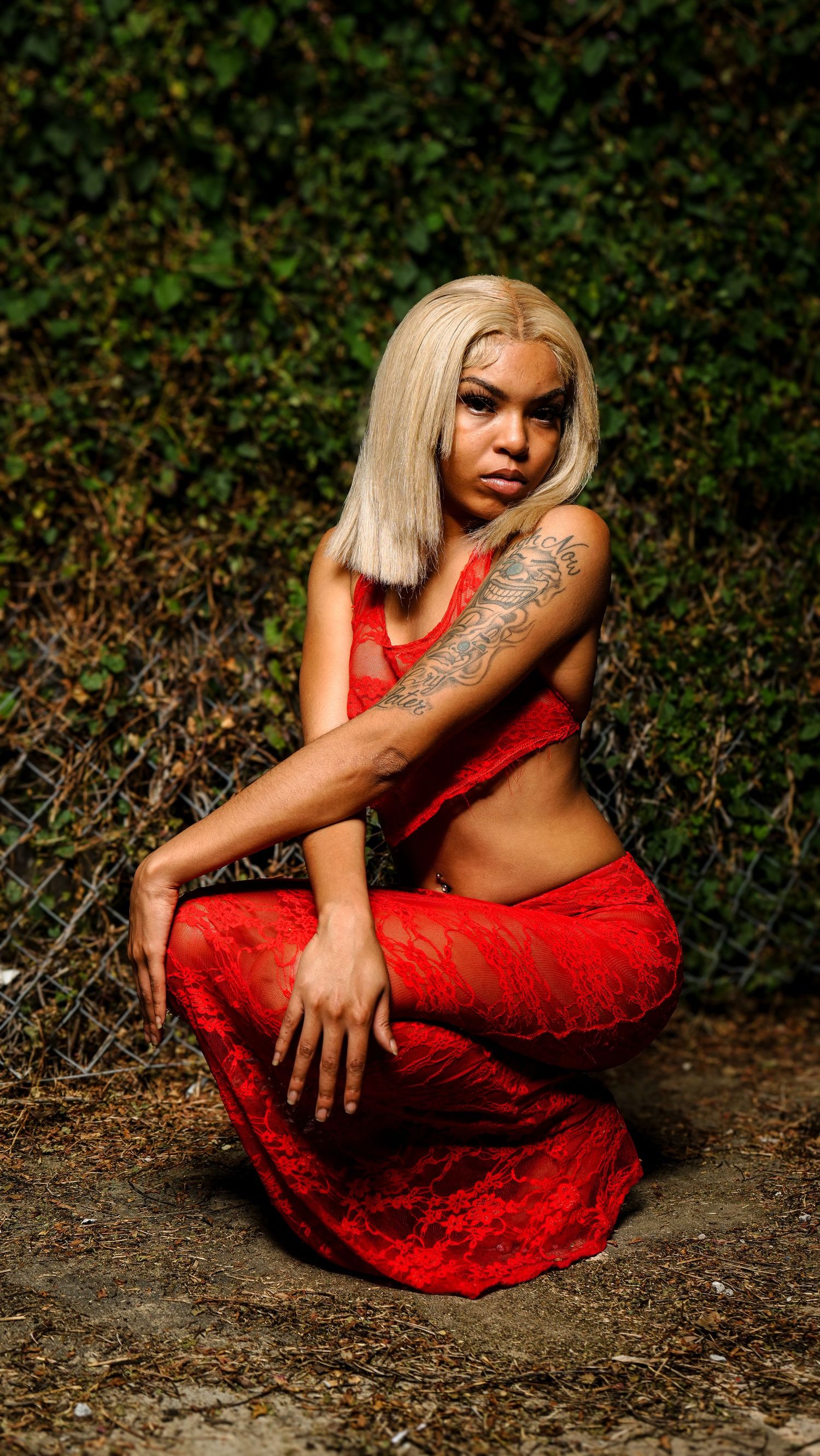Woman in red lace crop top and pants crouching outdoors with blonde hair and arm tattoo.