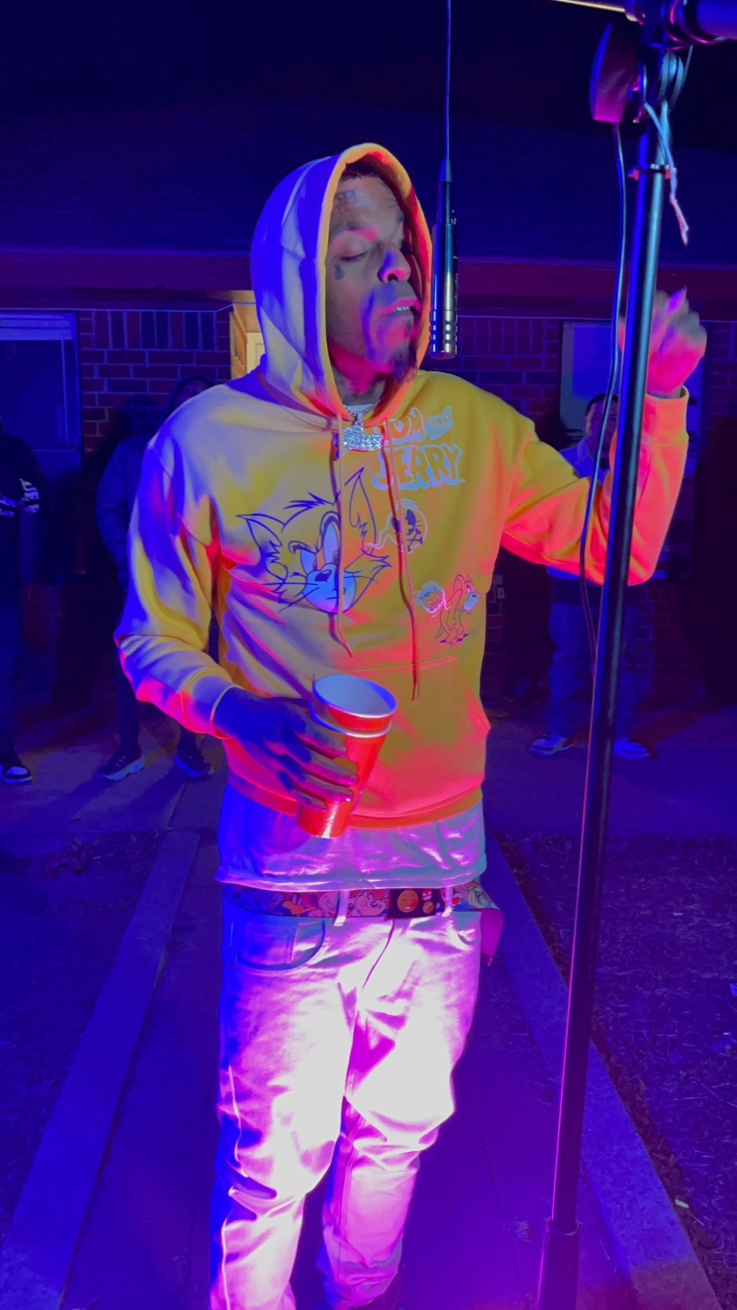 Person wearing yellow hoodie and white pants stands in colorful neon lighting creating vibrant purple and blue shadows.