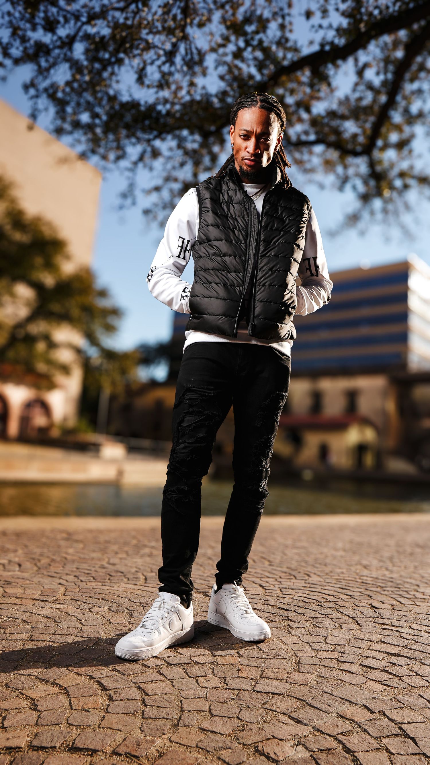 Young man in black puffer vest, white graphic long sleeve, black jeans, and white sneakers posing on cobblestone plaza.