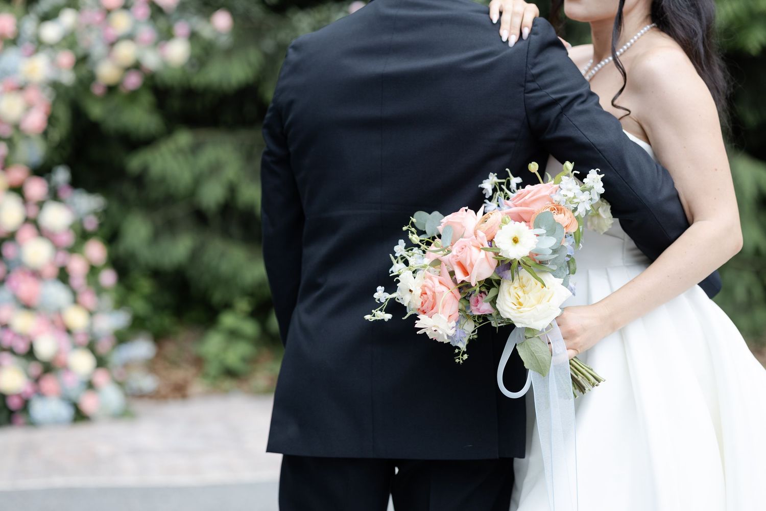 Couple embracing at wedding, bride holding pink and white bouquet with roses, groom in black suit, floral backdrop.