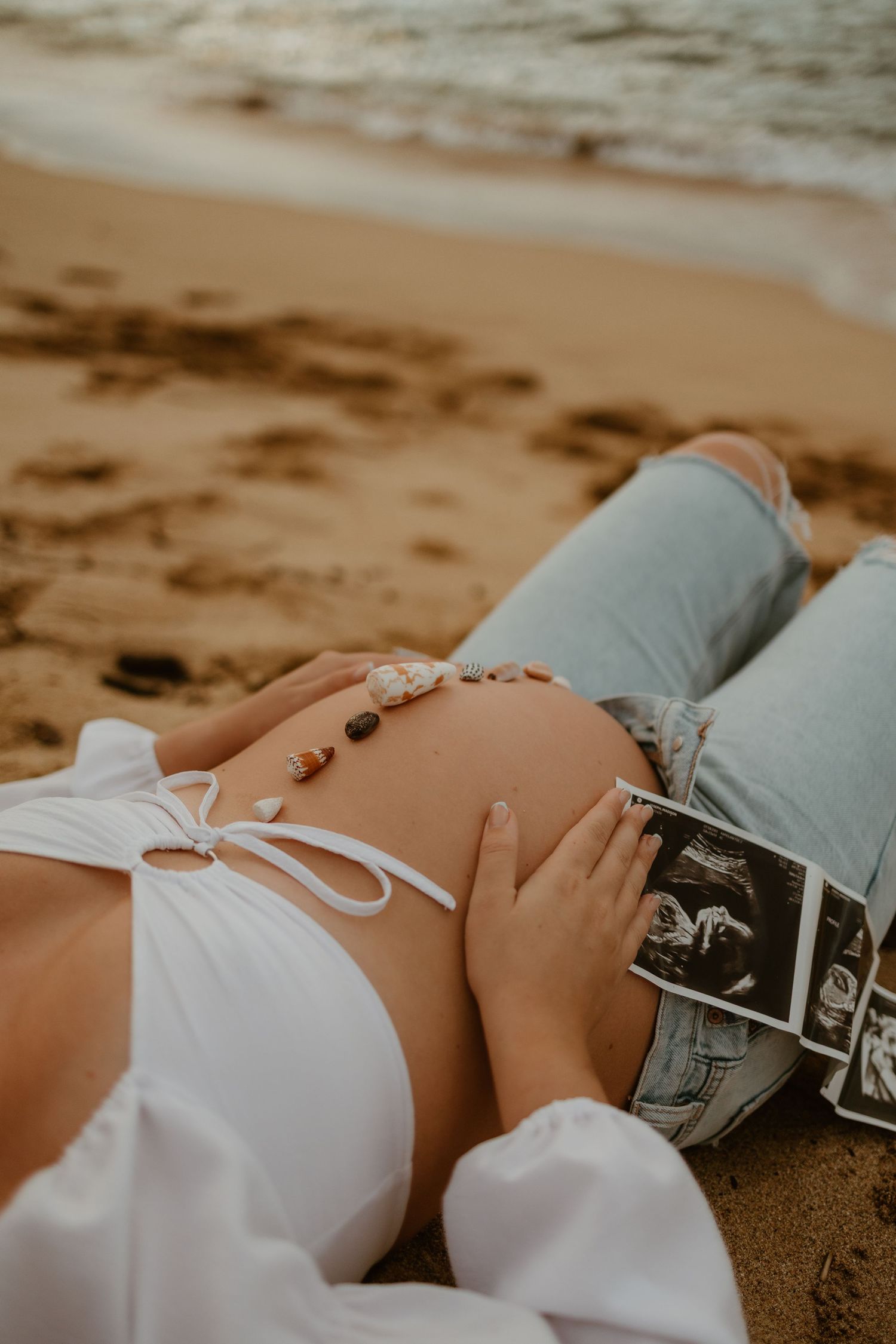 Pregnant woman in white top and jeans lying on a sandy beach with seashells on her belly and ultrasound photos beside her.