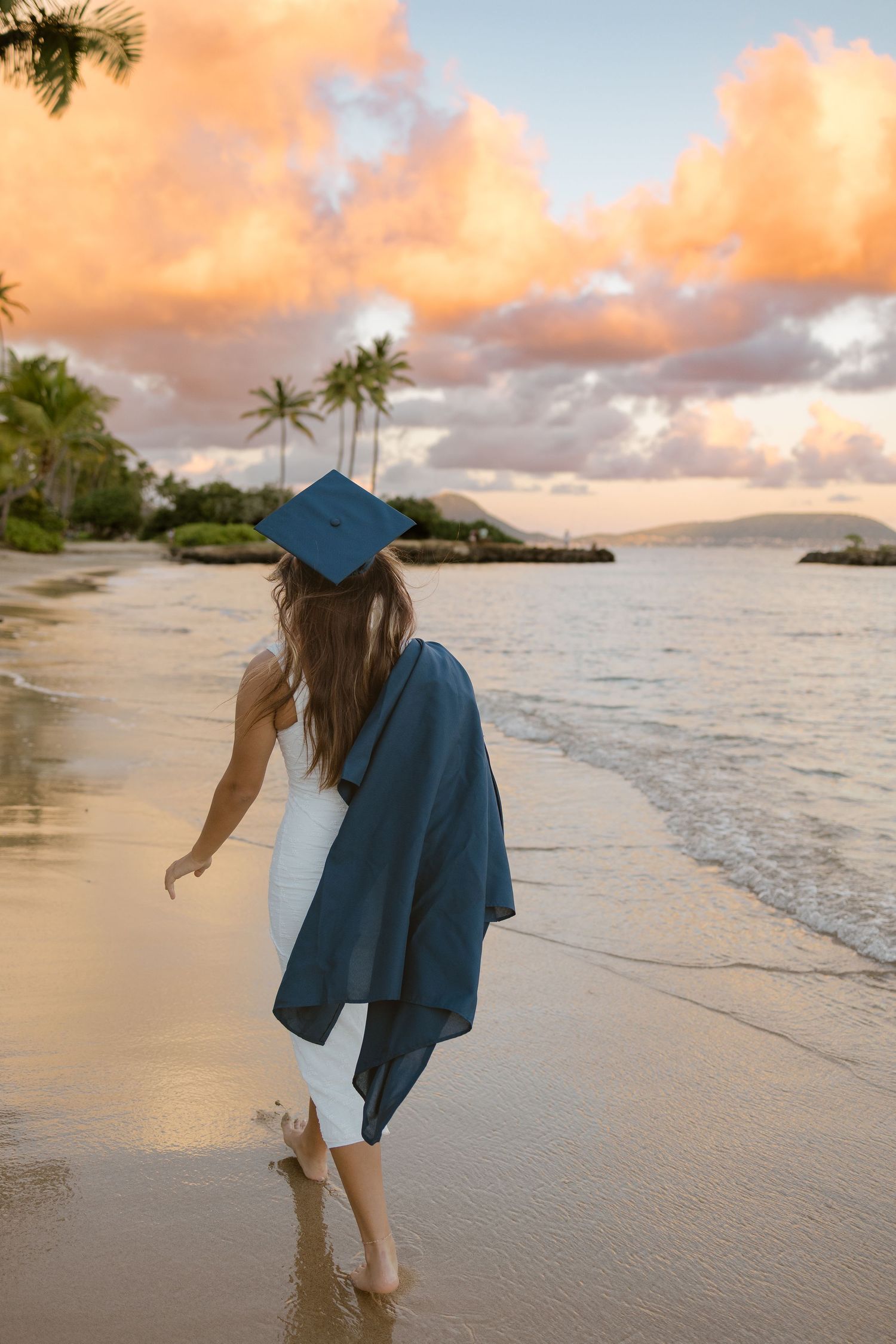 Graduate in a blue cap and gown walking barefoot along a tropical beach at sunset with palm trees and pink clouds.