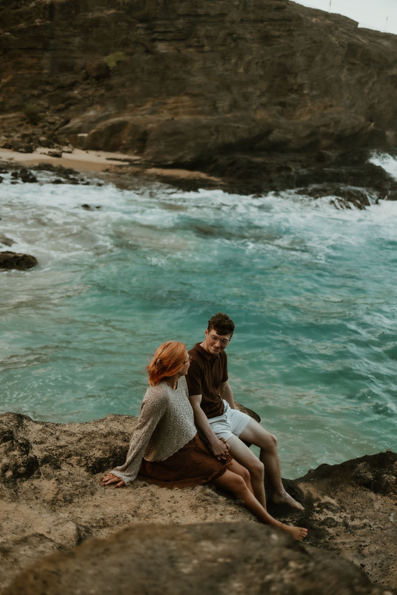 A couple sits close together on a rocky coastal ledge overlooking turquoise ocean waves crashing against rugged cliffs.