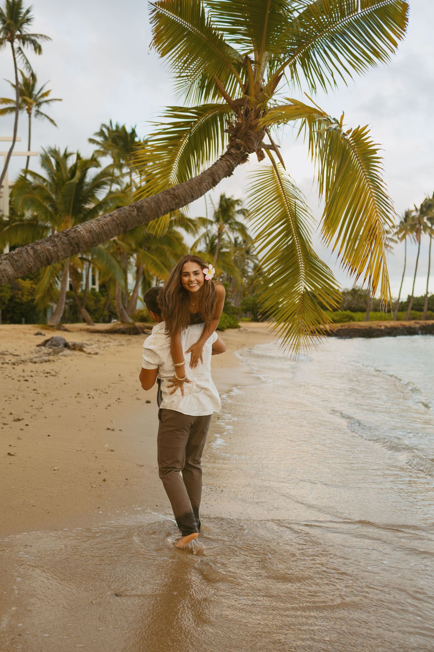 Smiling woman wearing a flower in her hair rides piggyback on a man along a tropical beach lined with palm trees.