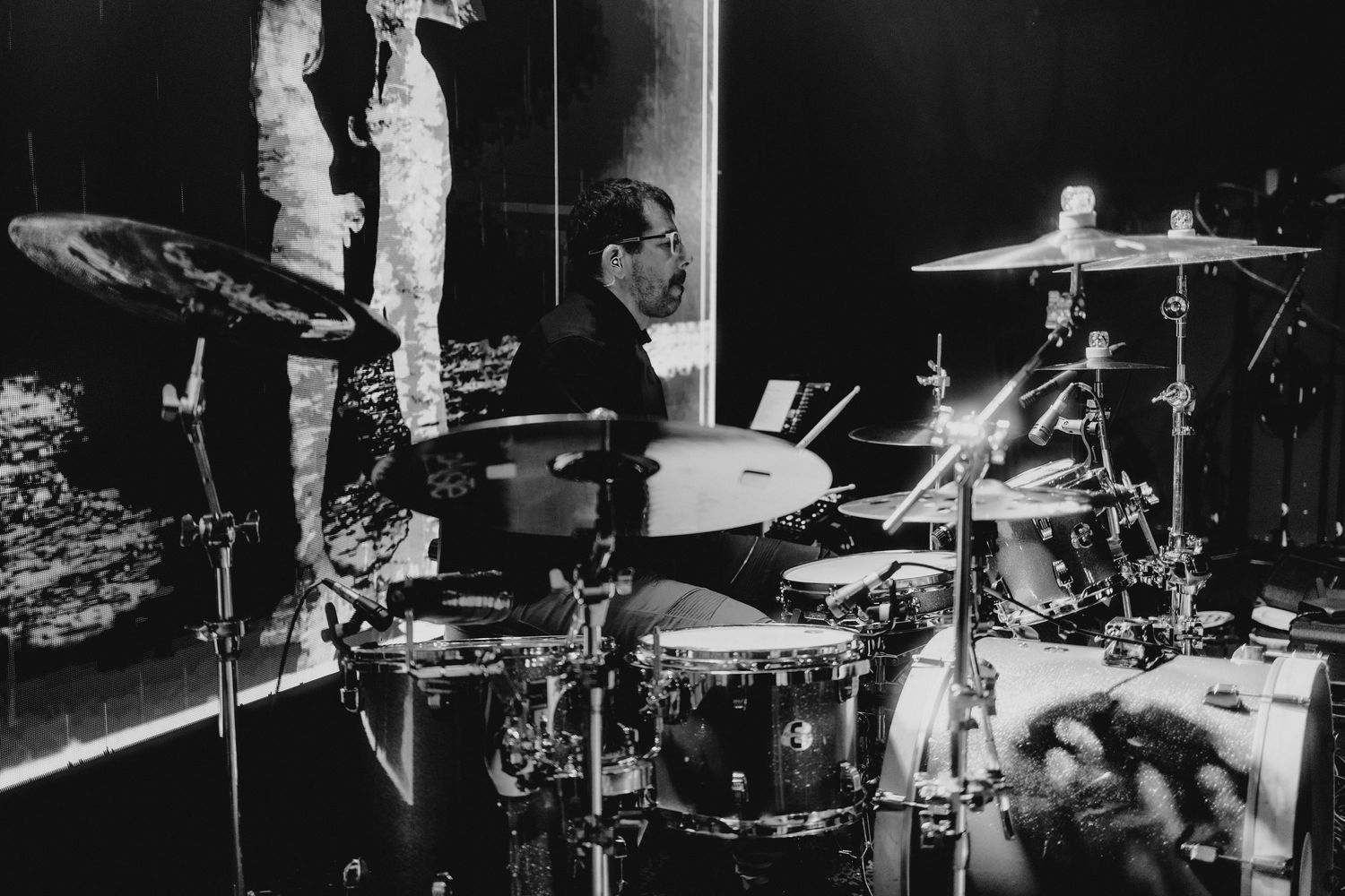 Black and white photo of a drummer performing on stage with cymbals and drum kit under stage lighting.