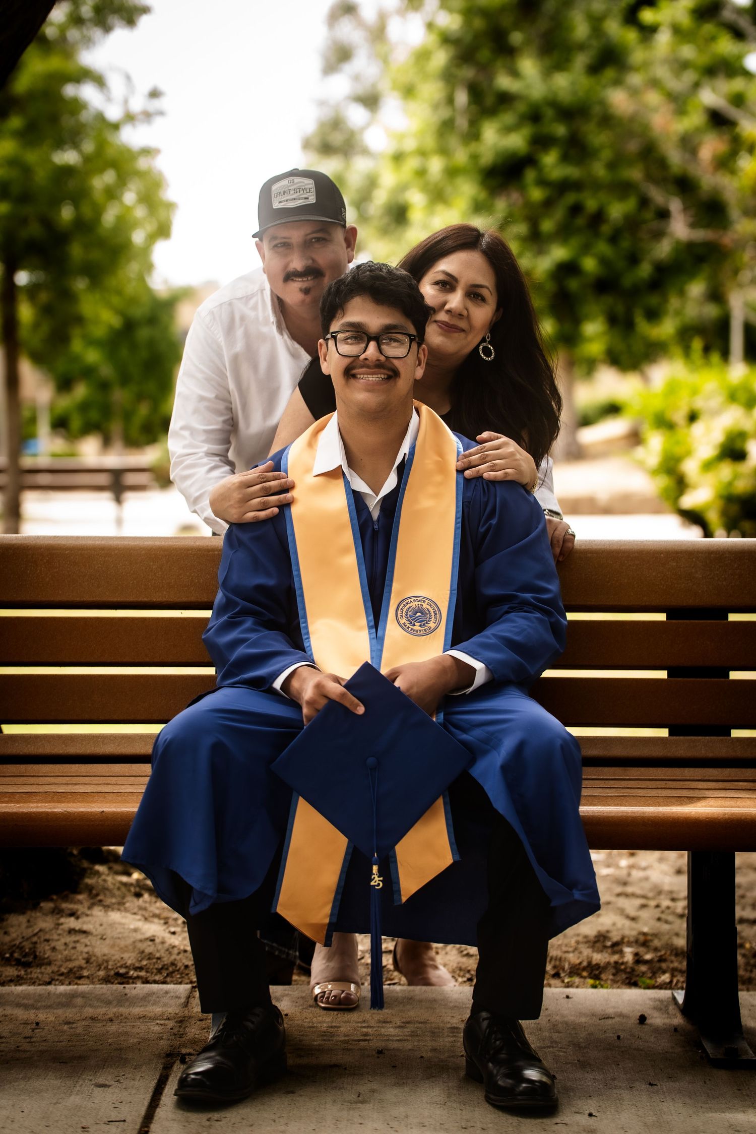 A proud graduate in blue cap and gown sitting on a bench with smiling parents behind him.