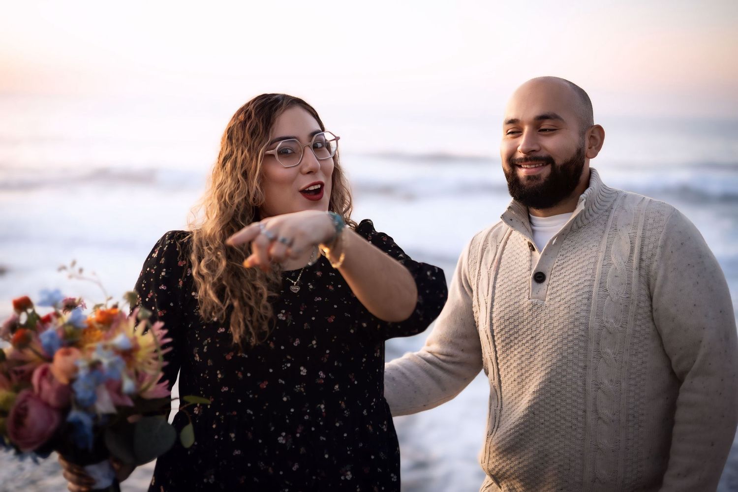 A smiling couple stands together near the ocean at sunset, the woman pointing excitedly.