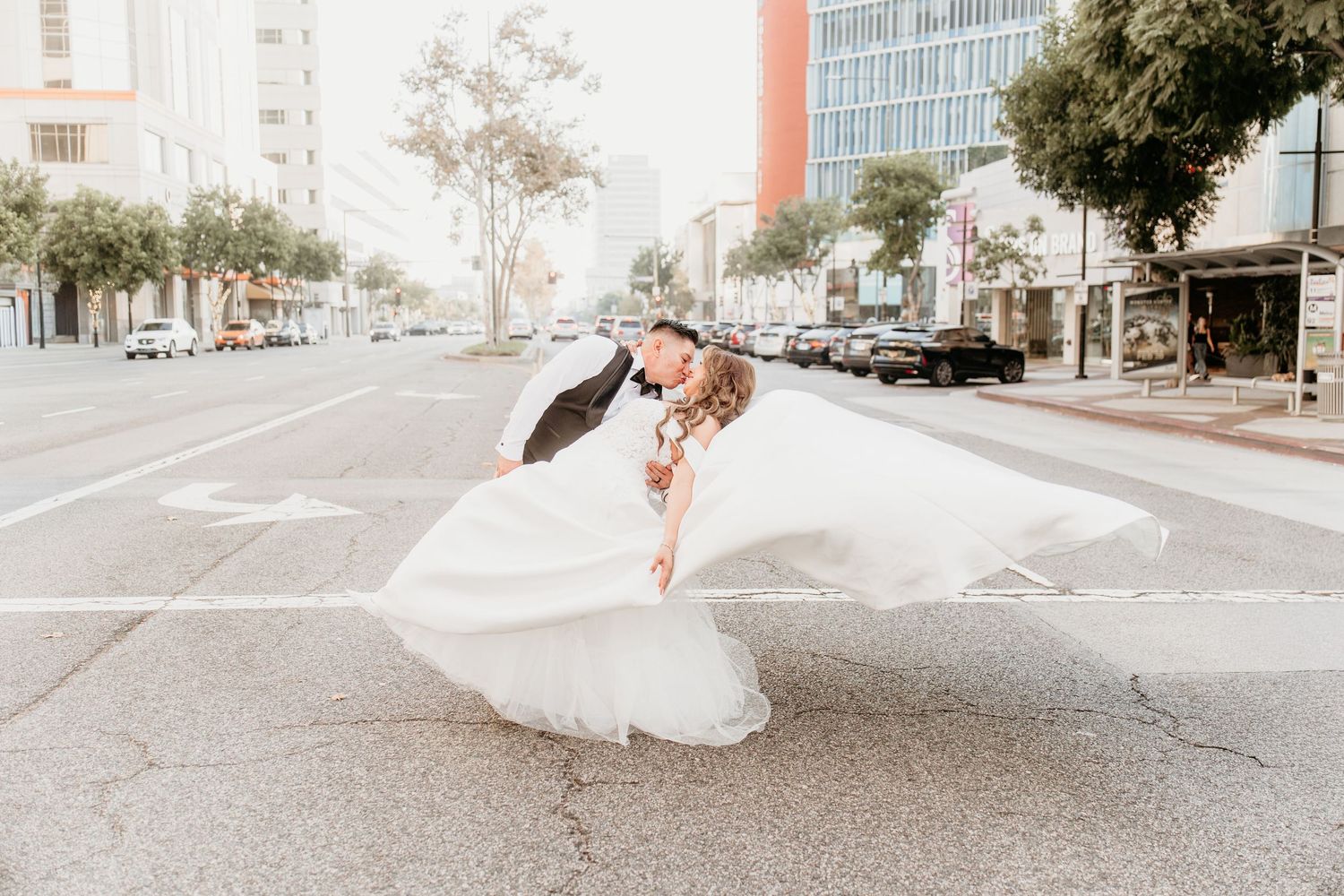 Groom dips bride in flowing white dress on urban city street intersection.